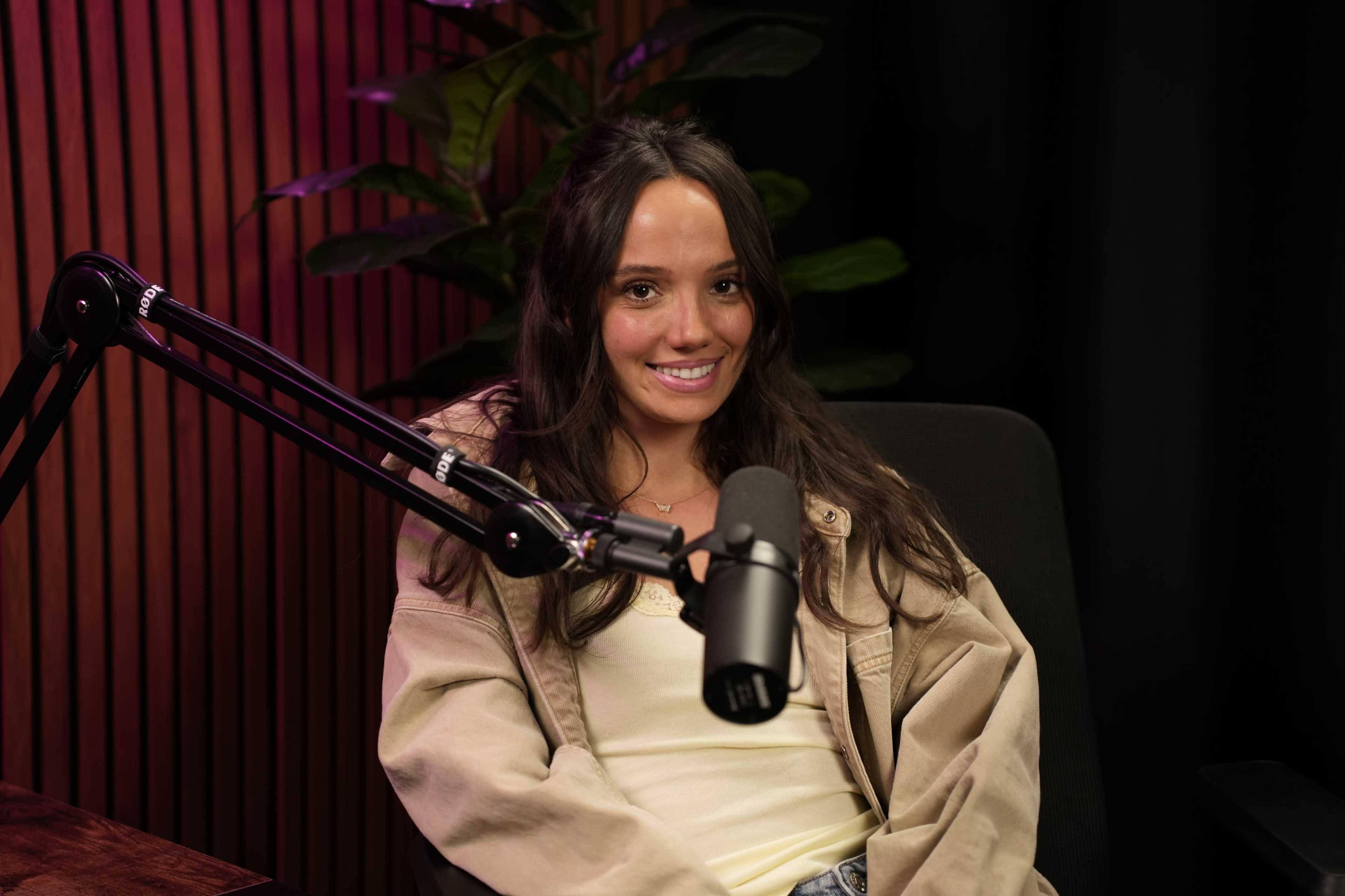 A young woman sits in a chair with a microphone in front of her against a backdrop of plants and wooden panels.