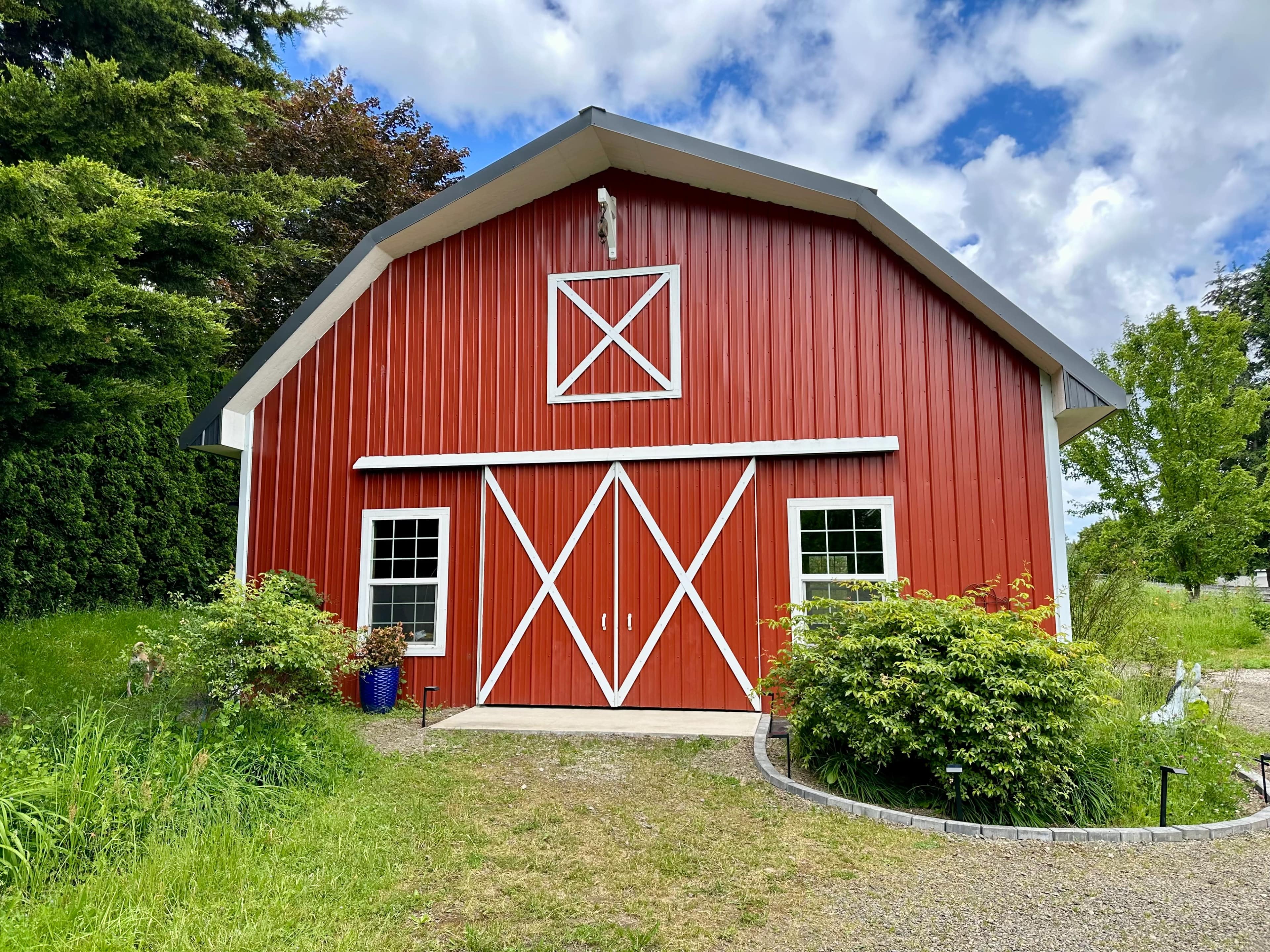 A red barn with a white trim and double doors, surrounded by green grass and trees under a partly cloudy sky.