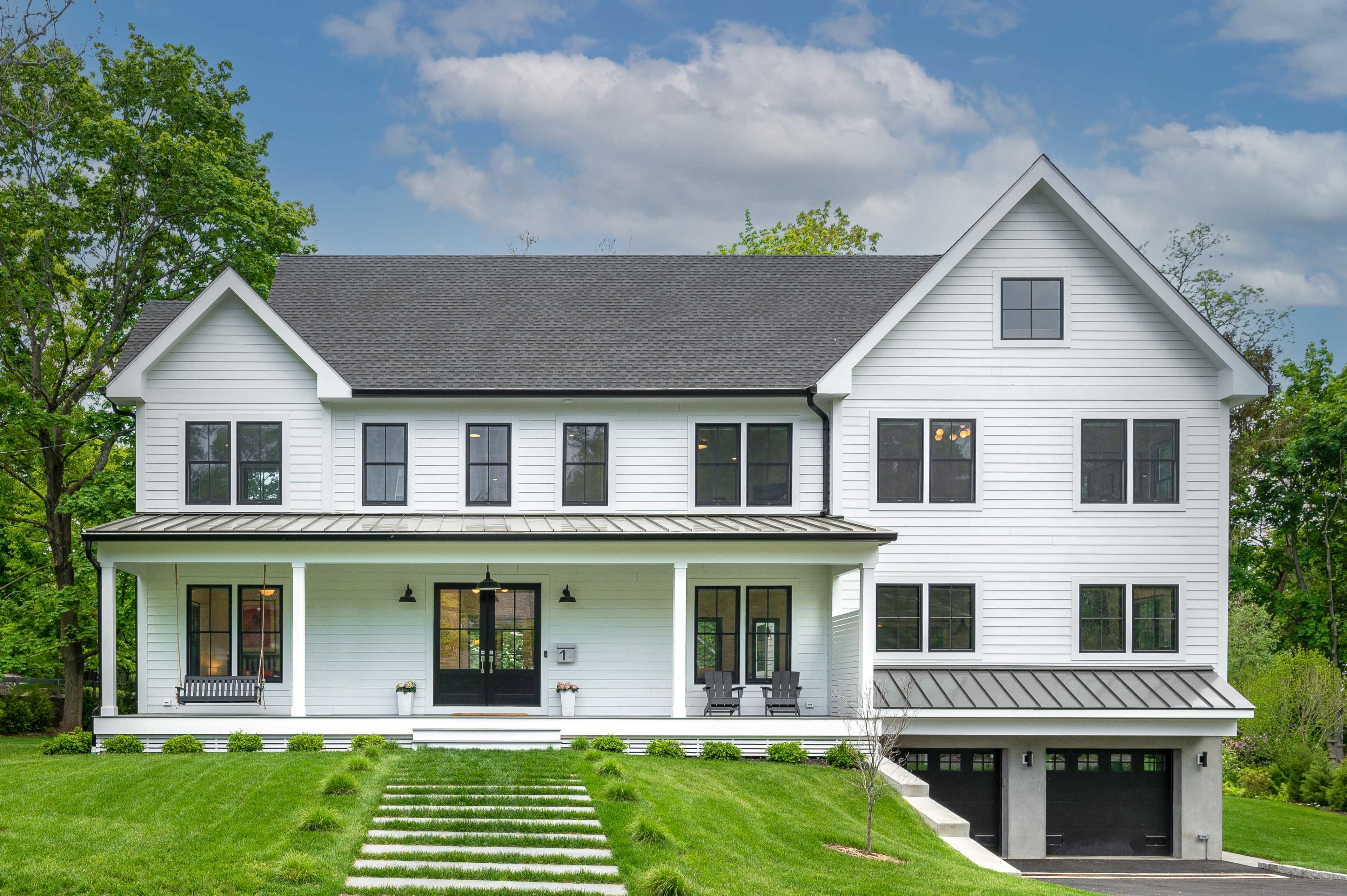 A large white house with a black roof and modern design, featuring a front porch and multiple windows, is set against a green landscape.