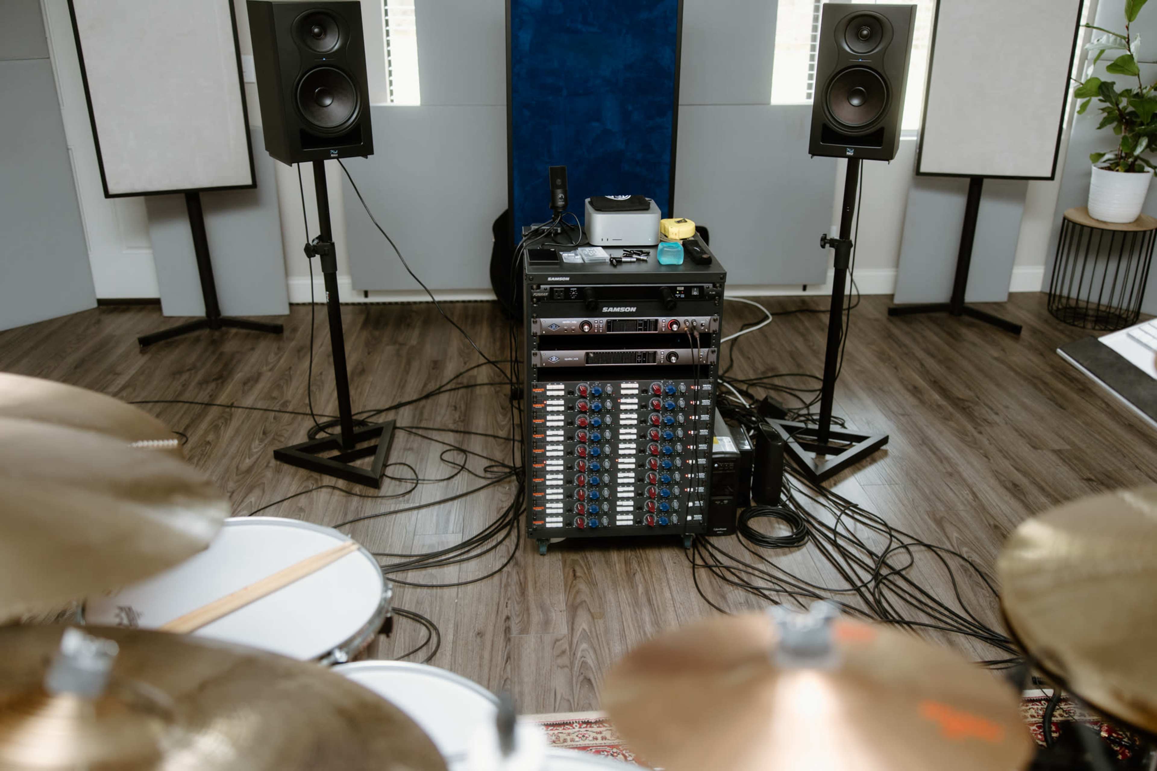 A sound mixing setup with a rack of audio equipment, two speakers on stands, and a drum kit in the foreground.
