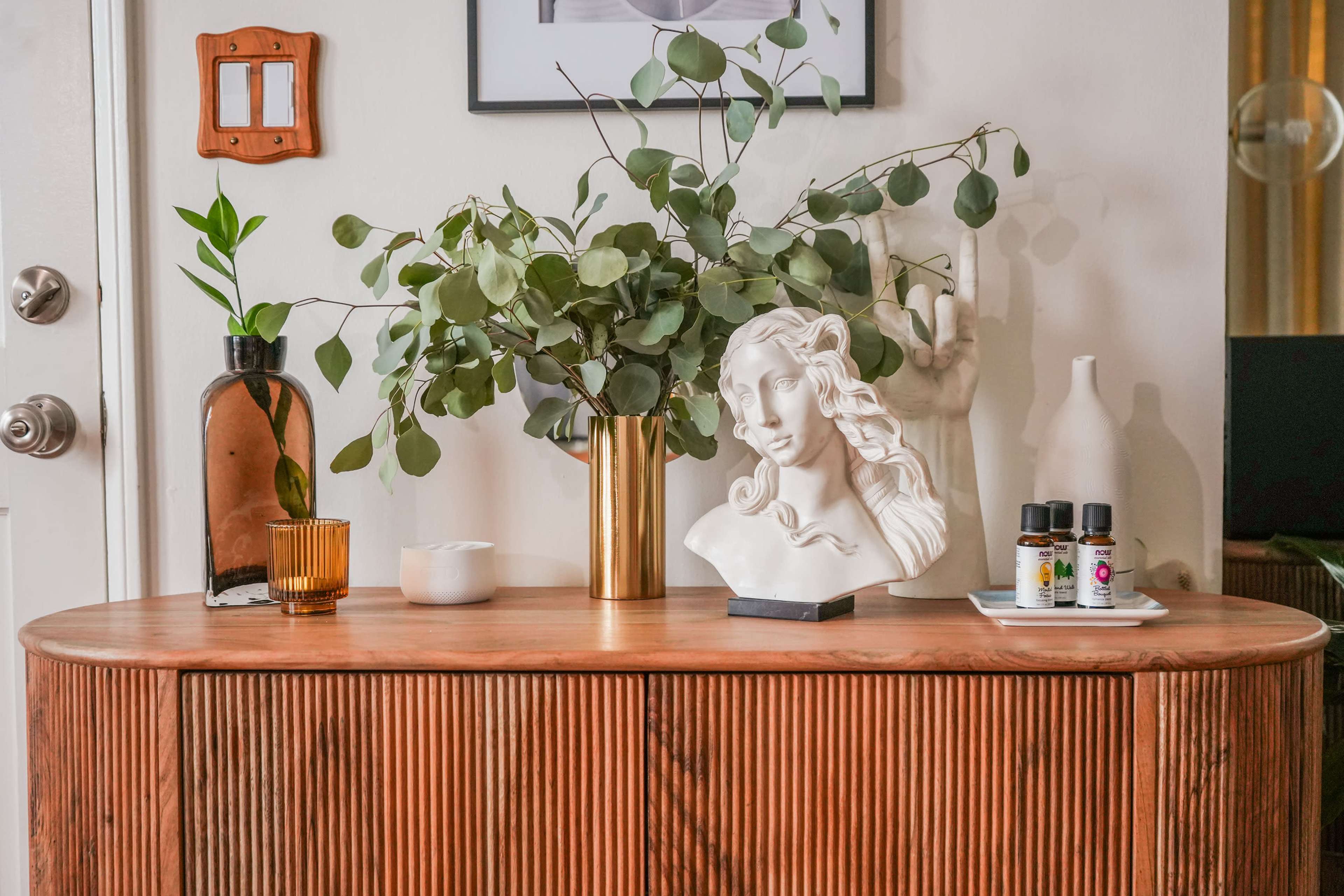 A wooden side table displays a white bust sculpture, a vase of greenery, a small plant, and bottles of essential oils.