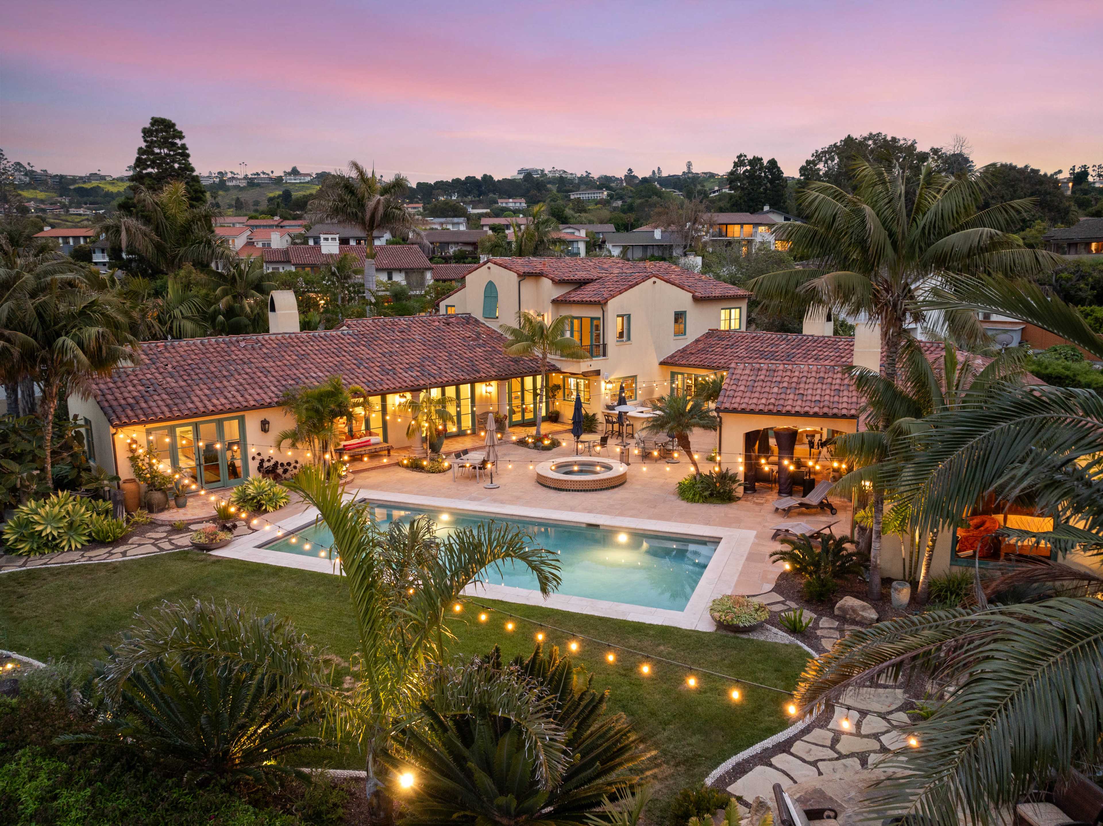 A luxurious home with a tiled roof surrounds a large pool and landscaped garden, set against a vibrant sunset sky.