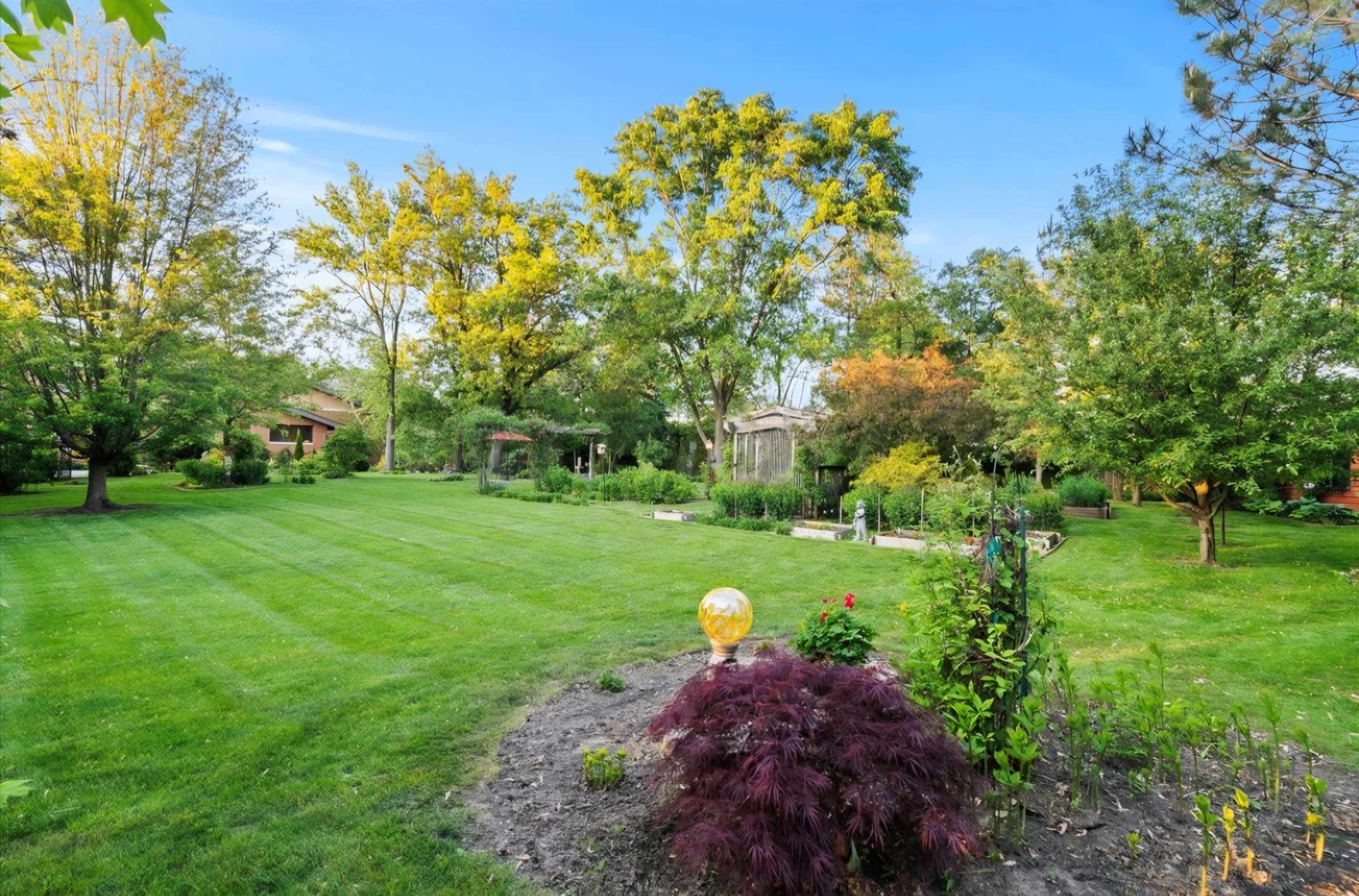 The image shows a large, well-manicured garden with various trees, shrubs, and a decorative orb in the foreground.