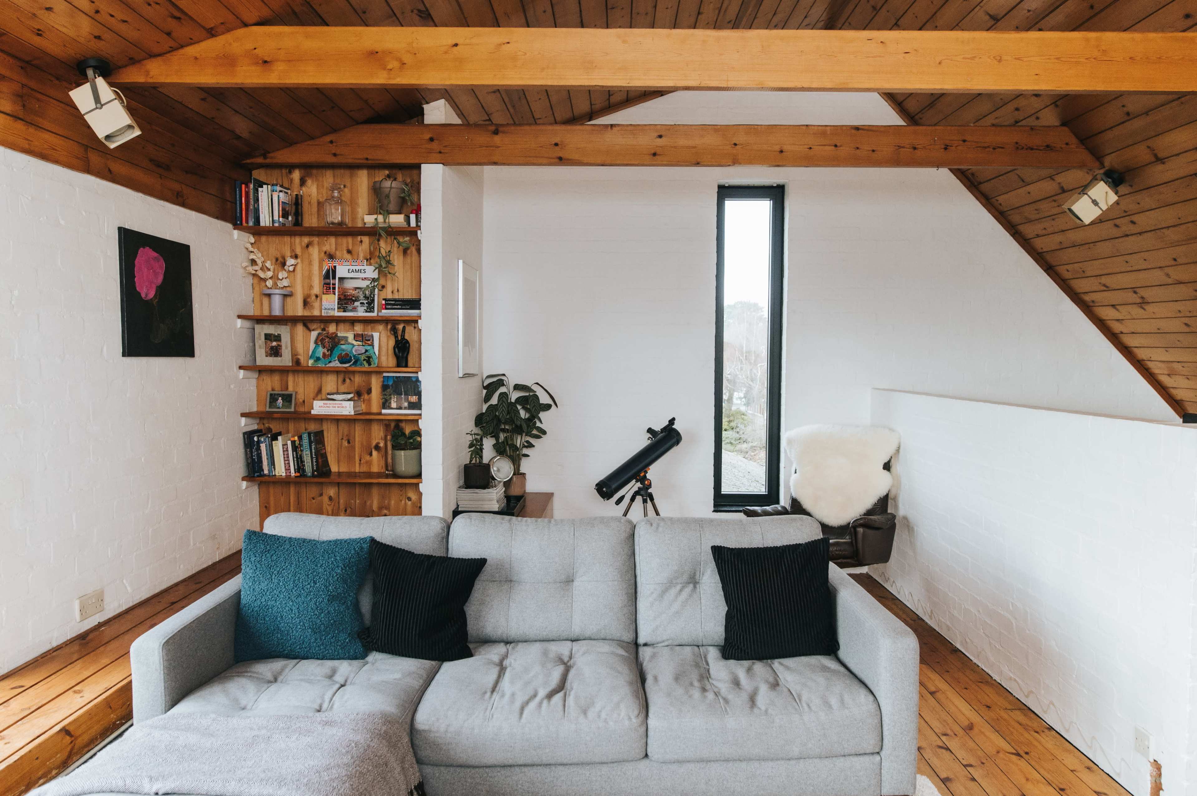 A cozy living room features a gray sofa positioned near a wooden beam ceiling, with a bookshelf and a tall window providing natural light.