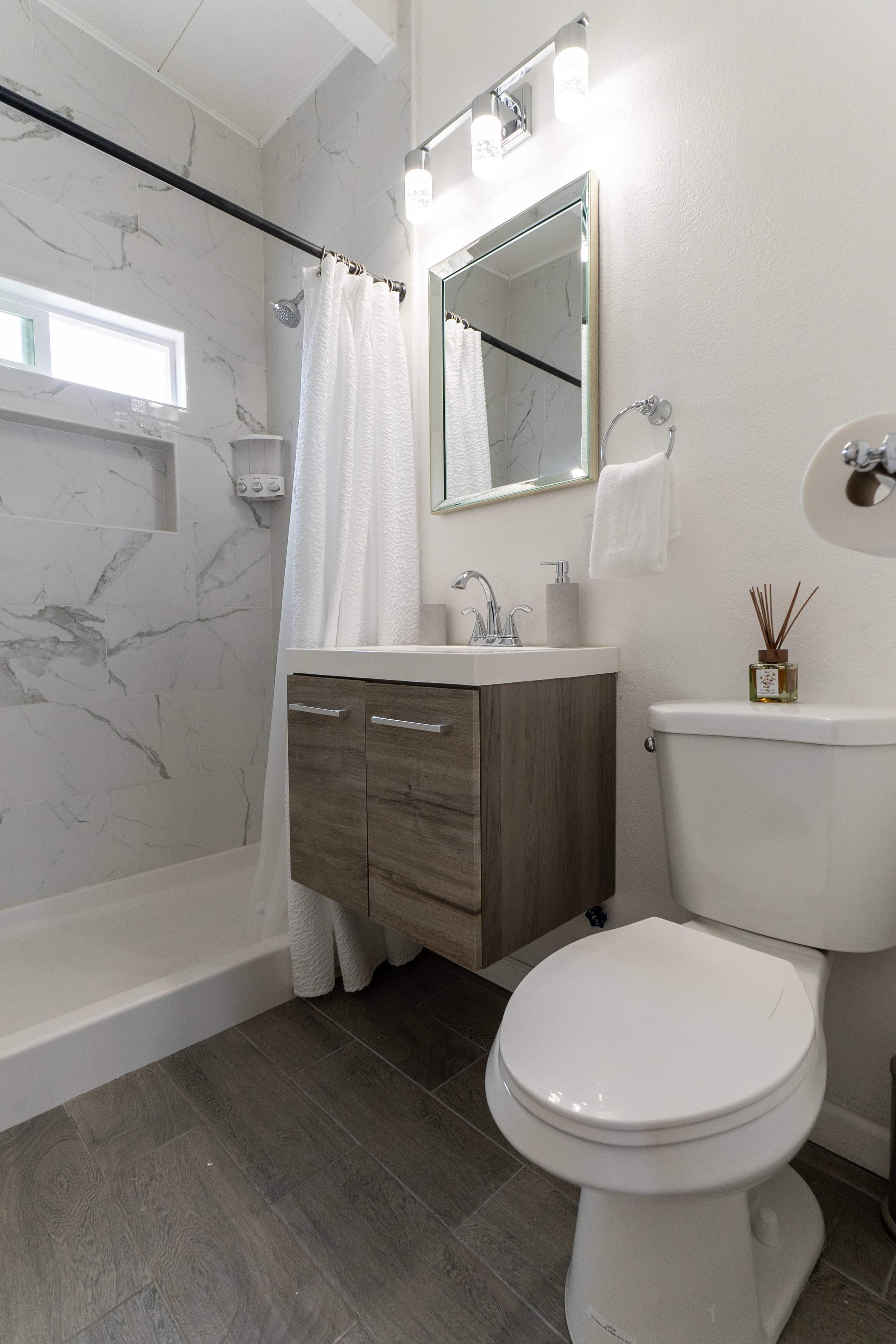 The image shows a modern bathroom featuring a shower, a vanity with wooden cabinets, a toilet, and white marble walls.