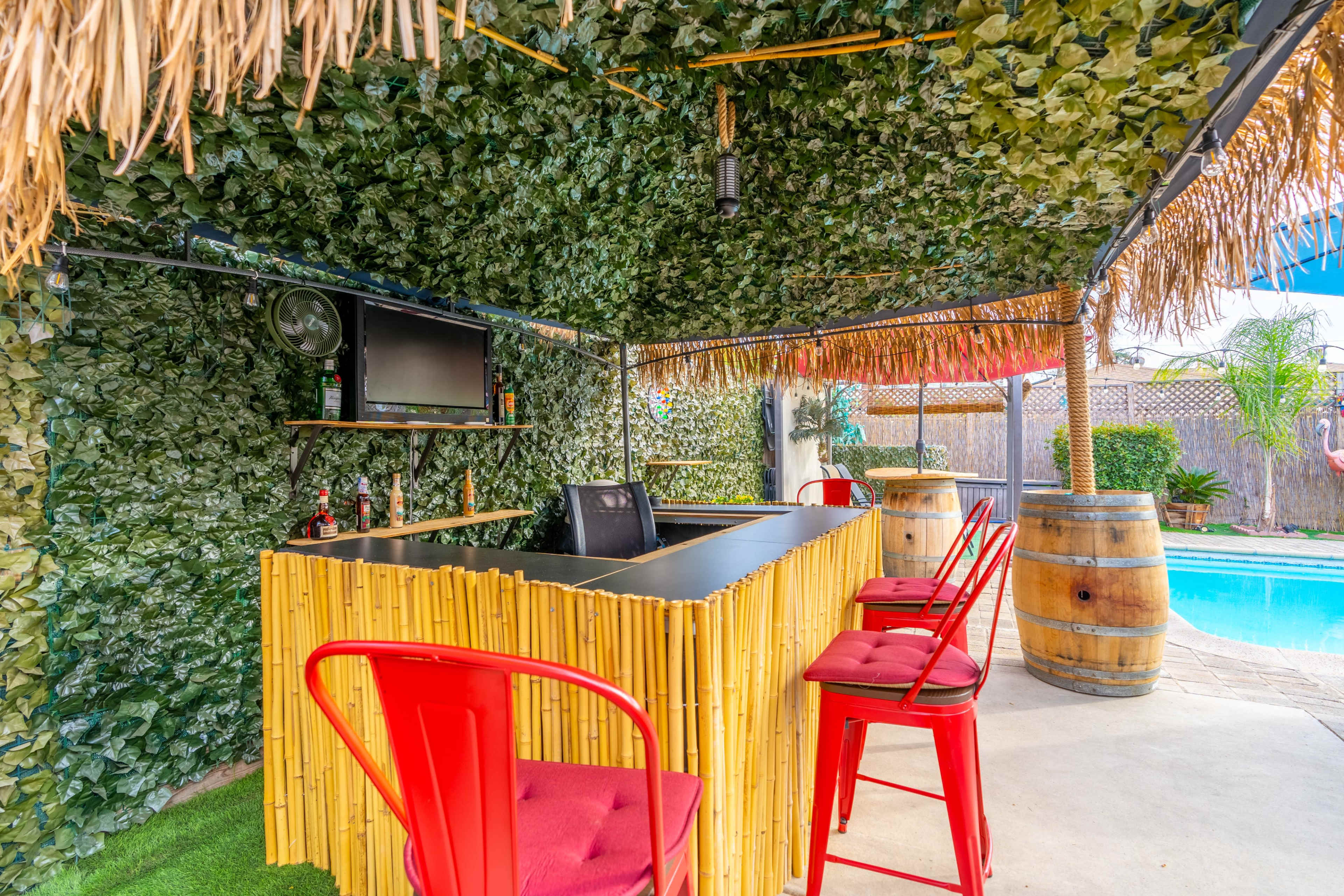 A backyard tiki bar with a thatched roof, bamboo counter, red metal stools, and a view of a swimming pool in the background.