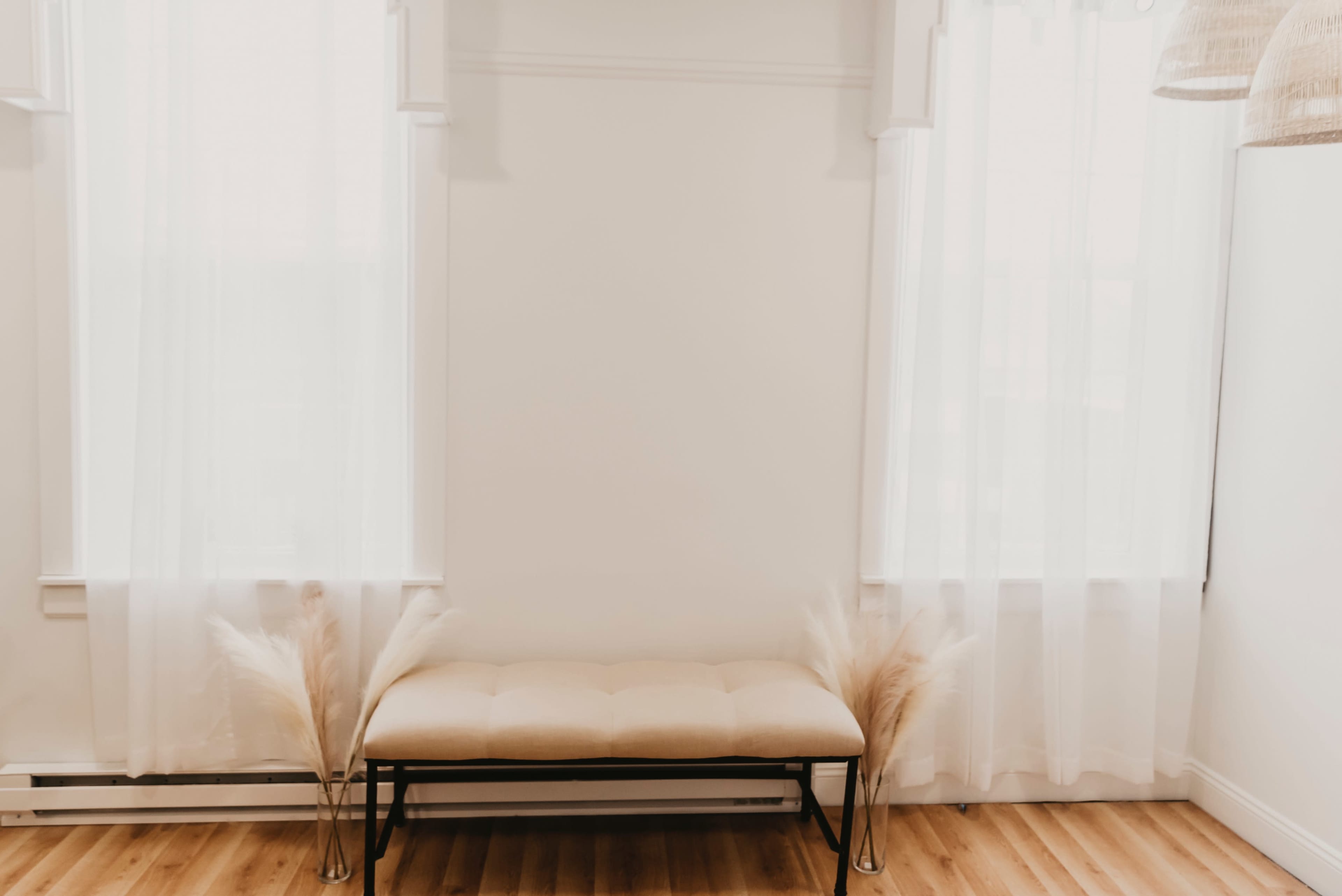 The image shows a simple, light-colored bench placed against a white wall with sheer curtains on either side and decorative dried grass arrangements.