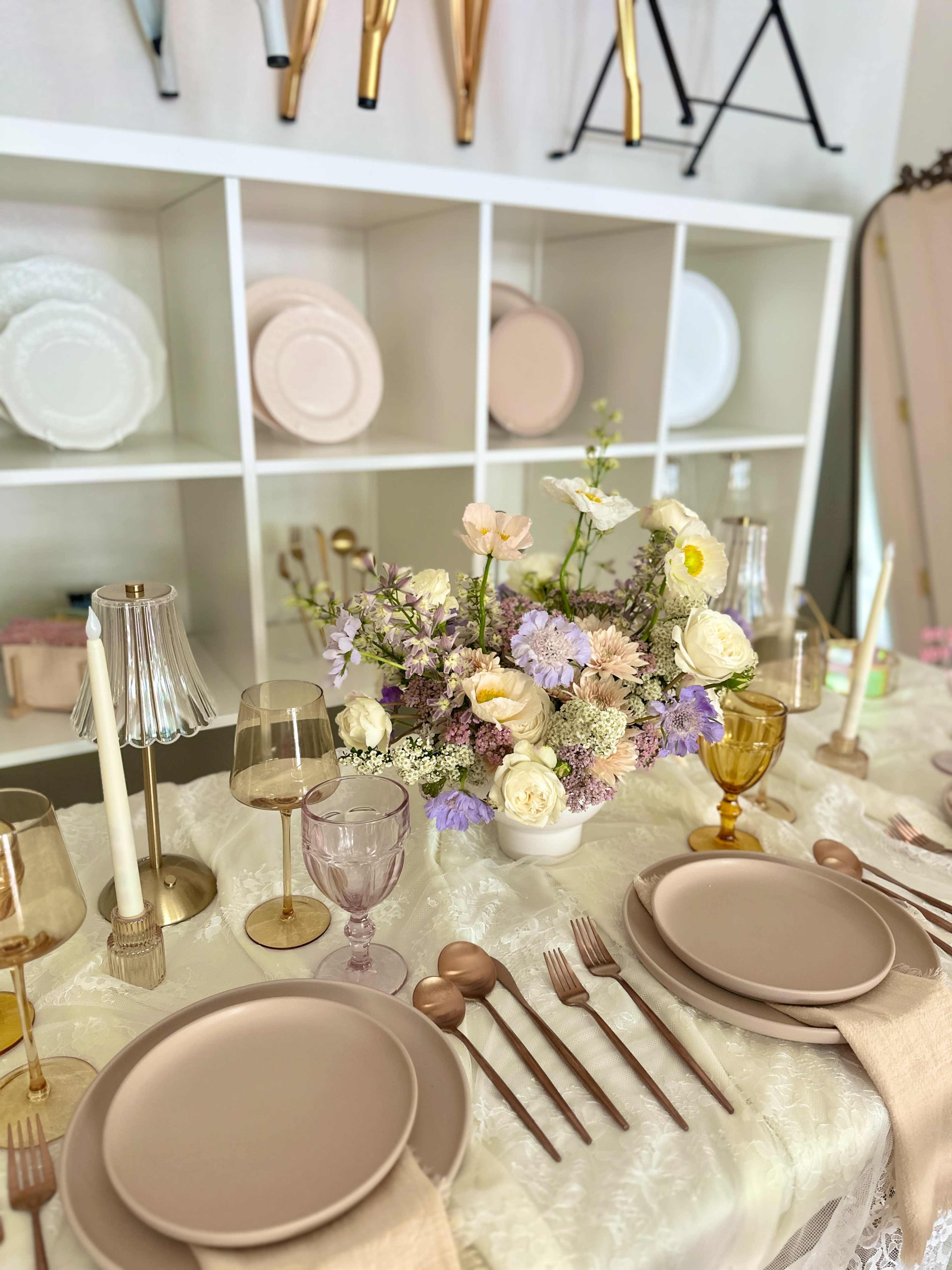 A dining table is elegantly arranged with pastel plates, glasses, and flatware, featuring a floral centerpiece amidst a white backdrop of shelves displaying various dishes.