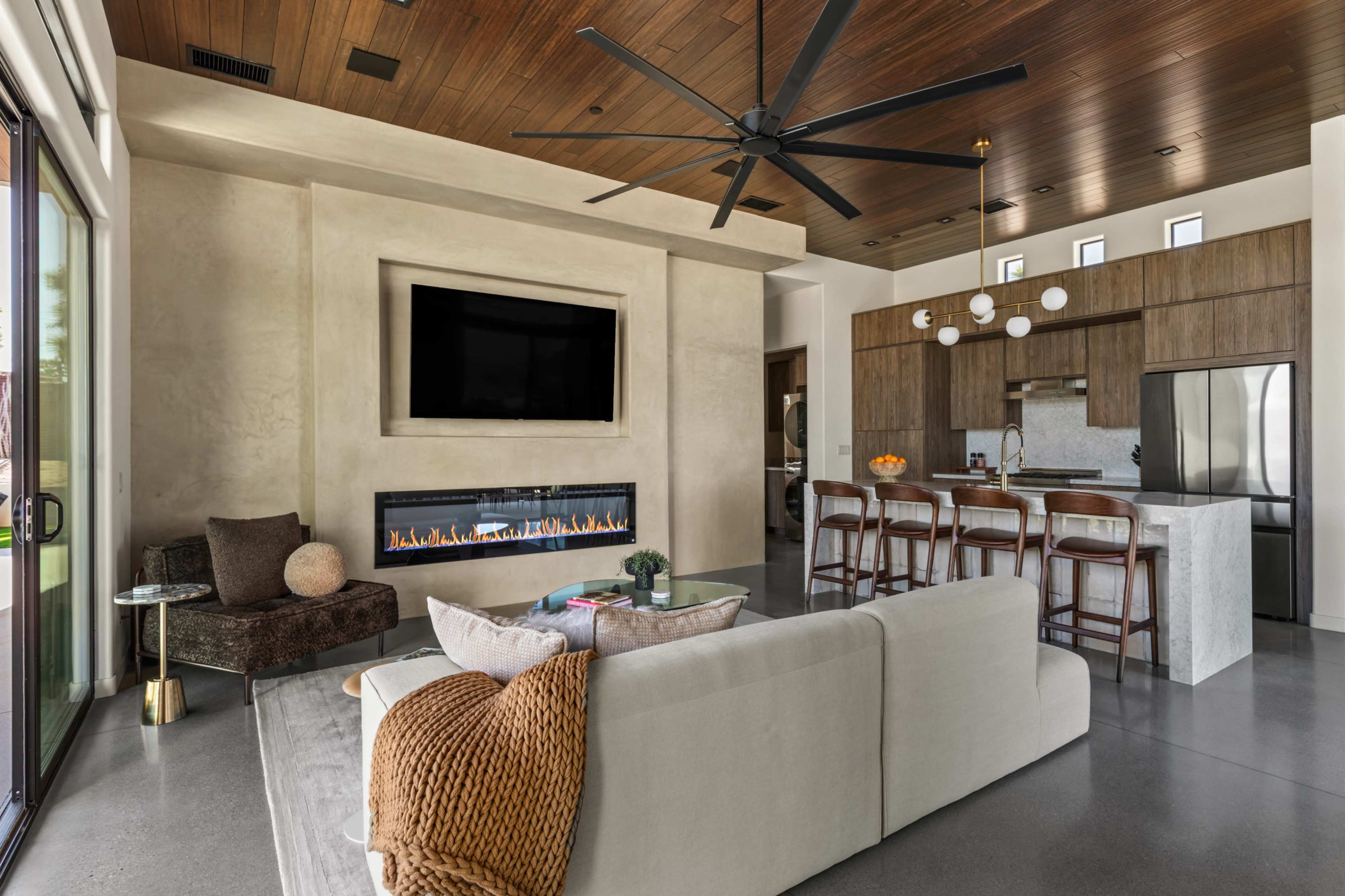 The image shows a modern living room with a large flat-screen TV above a linear fireplace, an open kitchen with bar seating, and a spacious seating area.