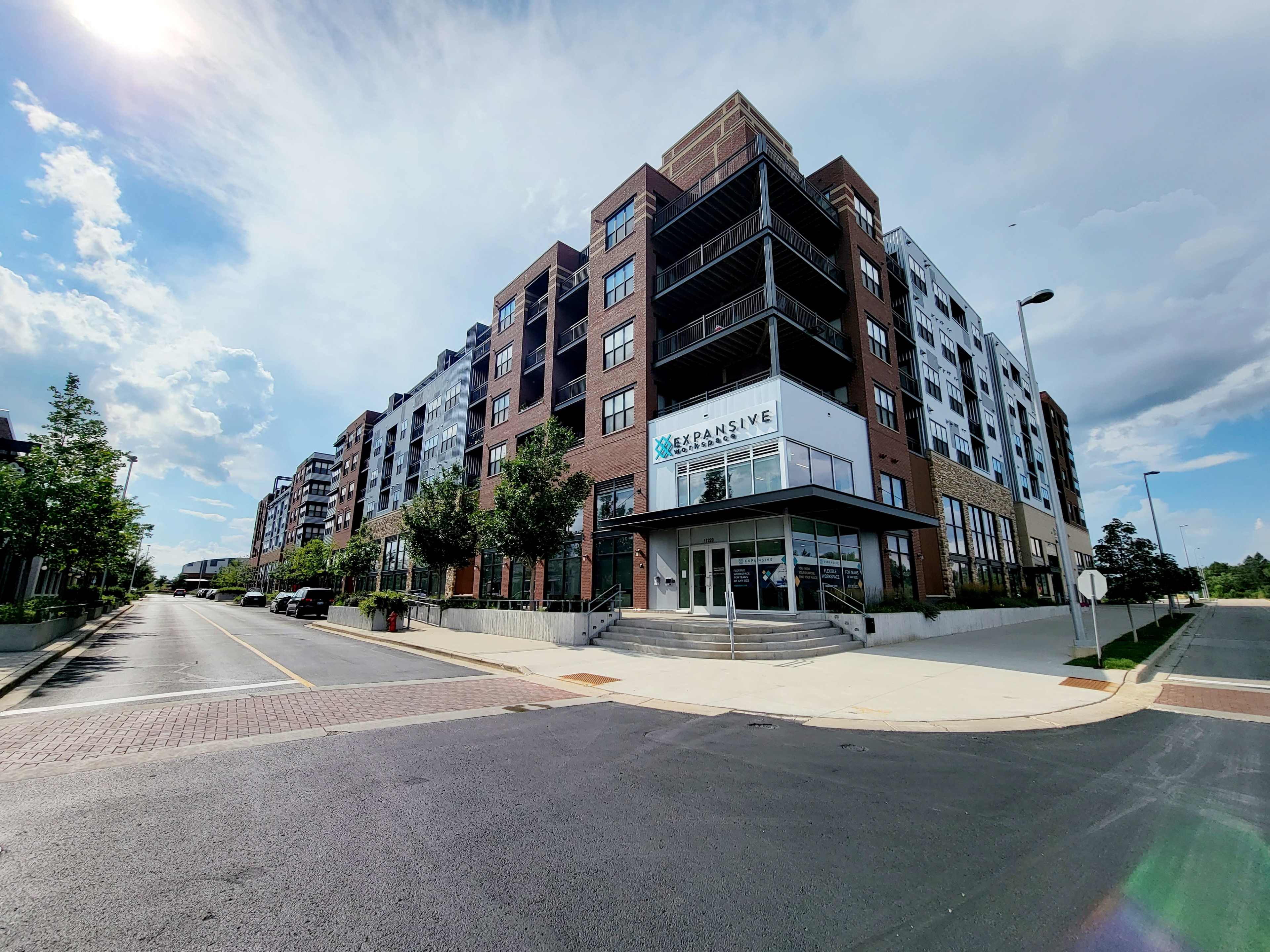 The image shows a modern multi-story apartment building situated at the corner of a street, with landscaped greenery in the foreground and a blue sky overhead.