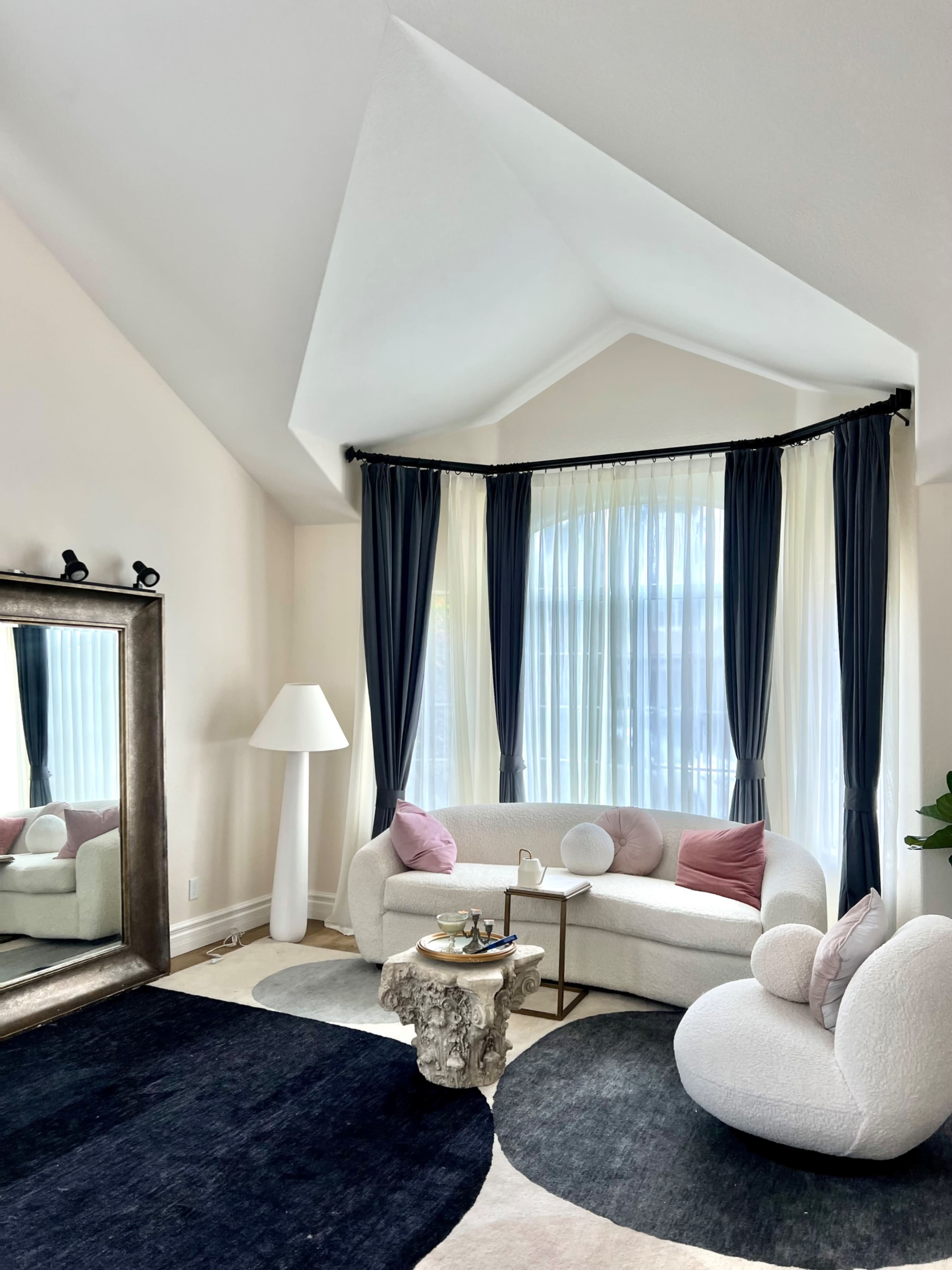 A modern living room with a curved sofa, a round coffee table, large windows with sheer curtains, and a tall floor lamp, all set on patterned rugs.