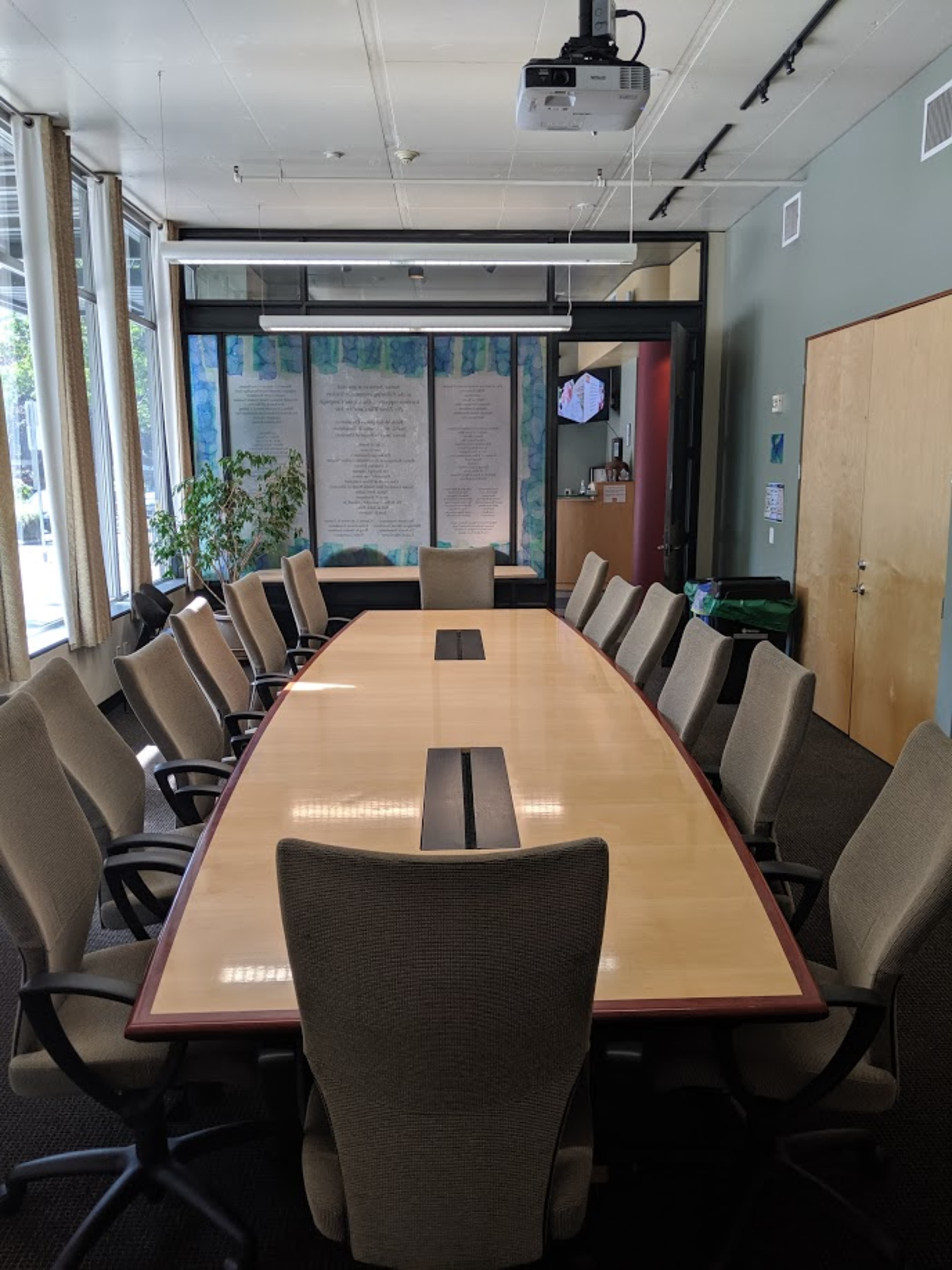 A large conference room features a rectangular table surrounded by gray office chairs, with a projector mounted on the ceiling and walls adorned with large windows.