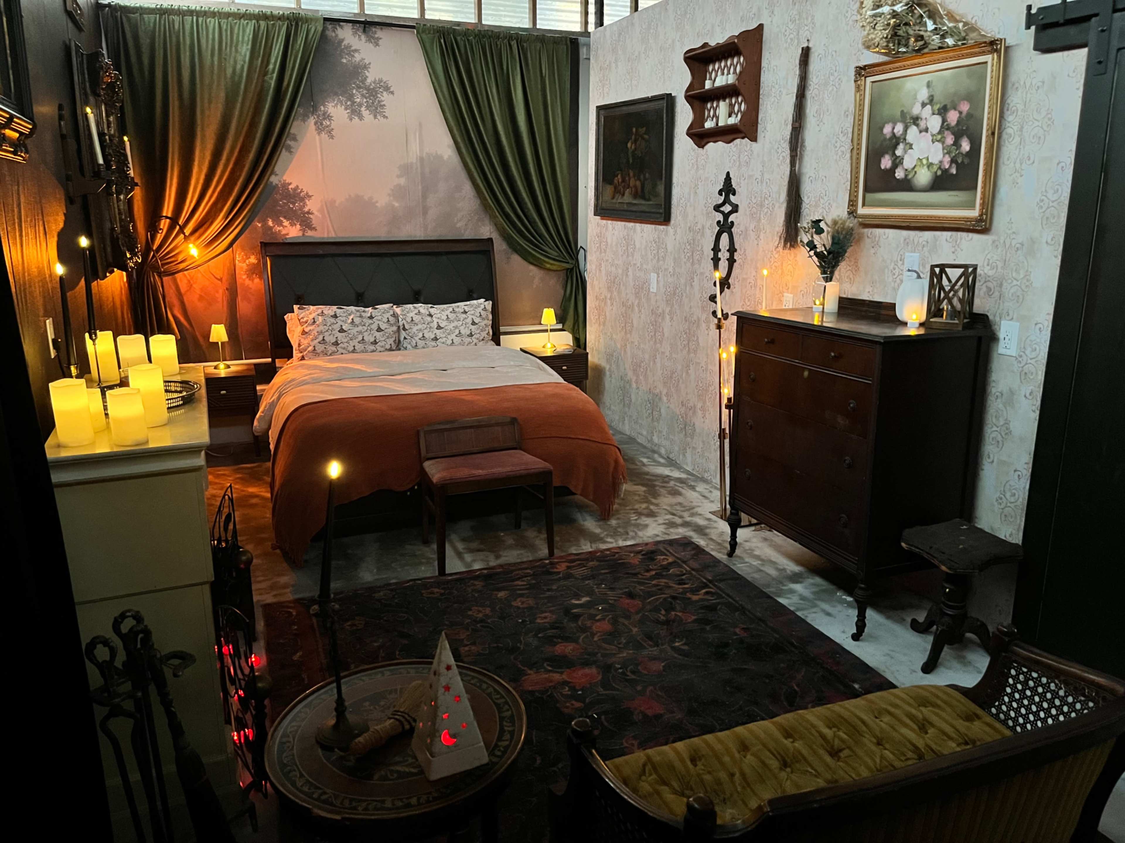 The image depicts a cozy bedroom with a bed, patterned curtains, a nightstand with candles, and various decorative elements, including paintings and a small seating area.