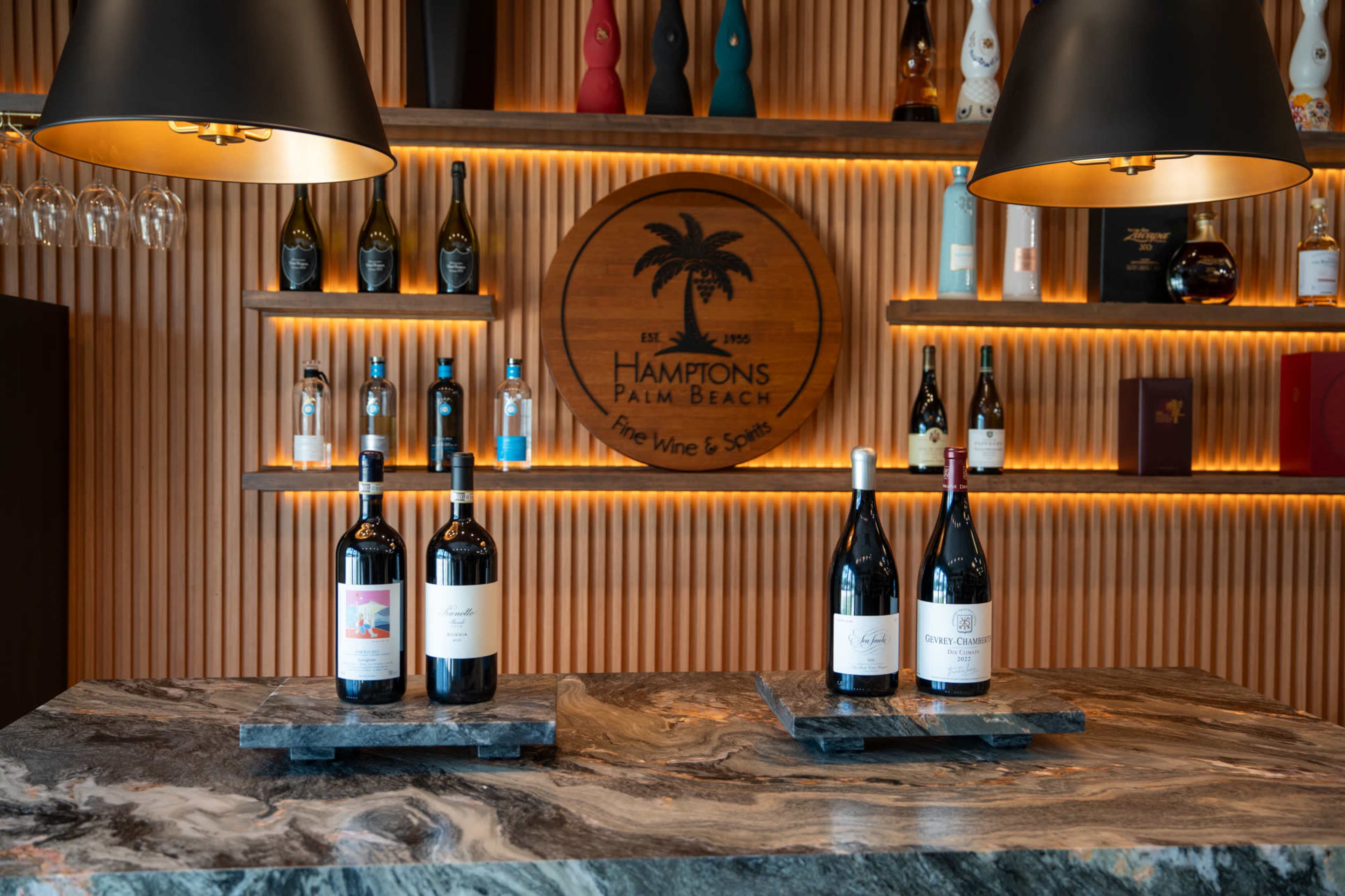 The image features a wine display with bottles arranged on marble surfaces against a wooden backdrop with a decorative sign for "Hamptons Palm Beach."