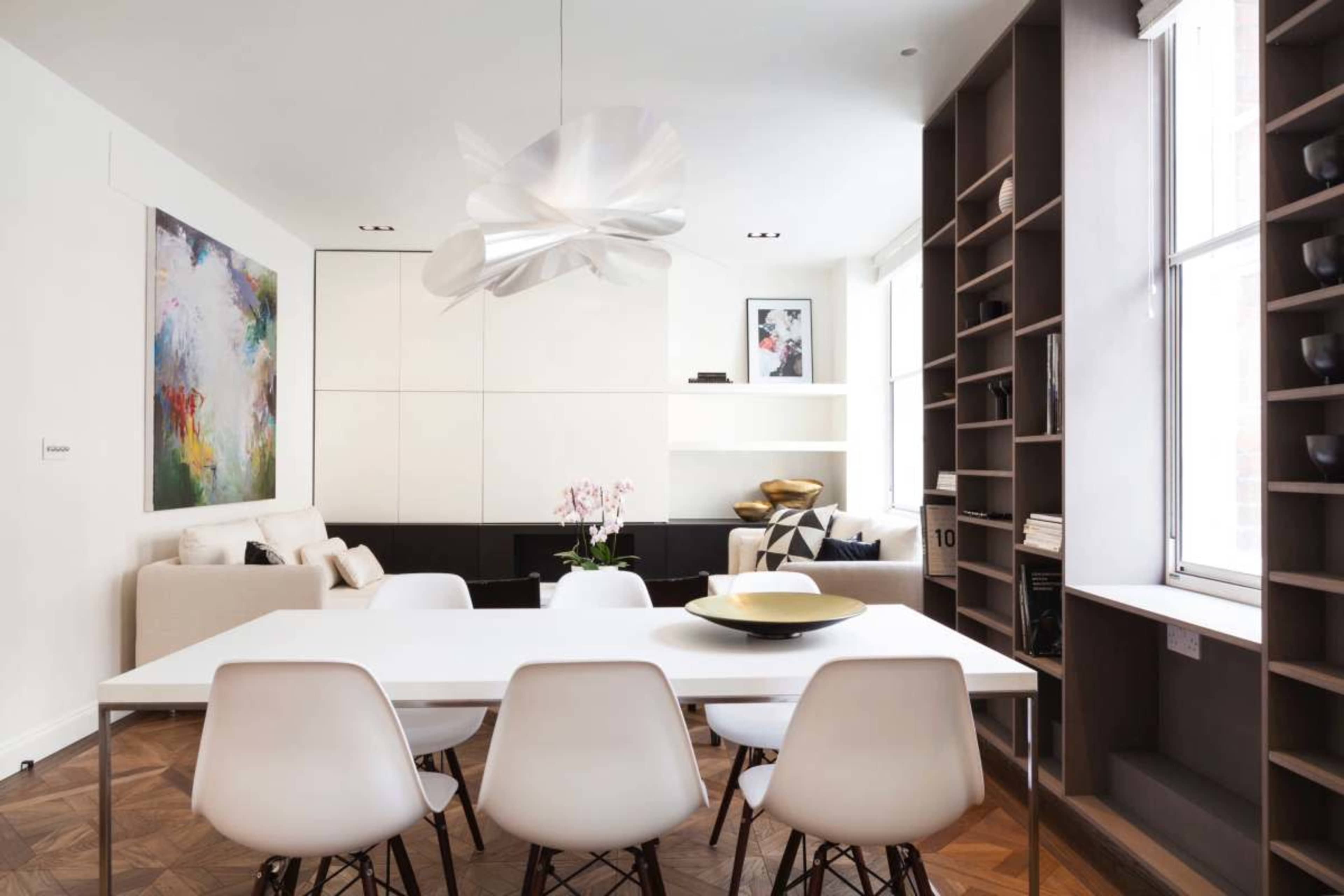 A modern dining area features a white table surrounded by six chairs, with a sleek light fixture above and a cozy seating area in the background.