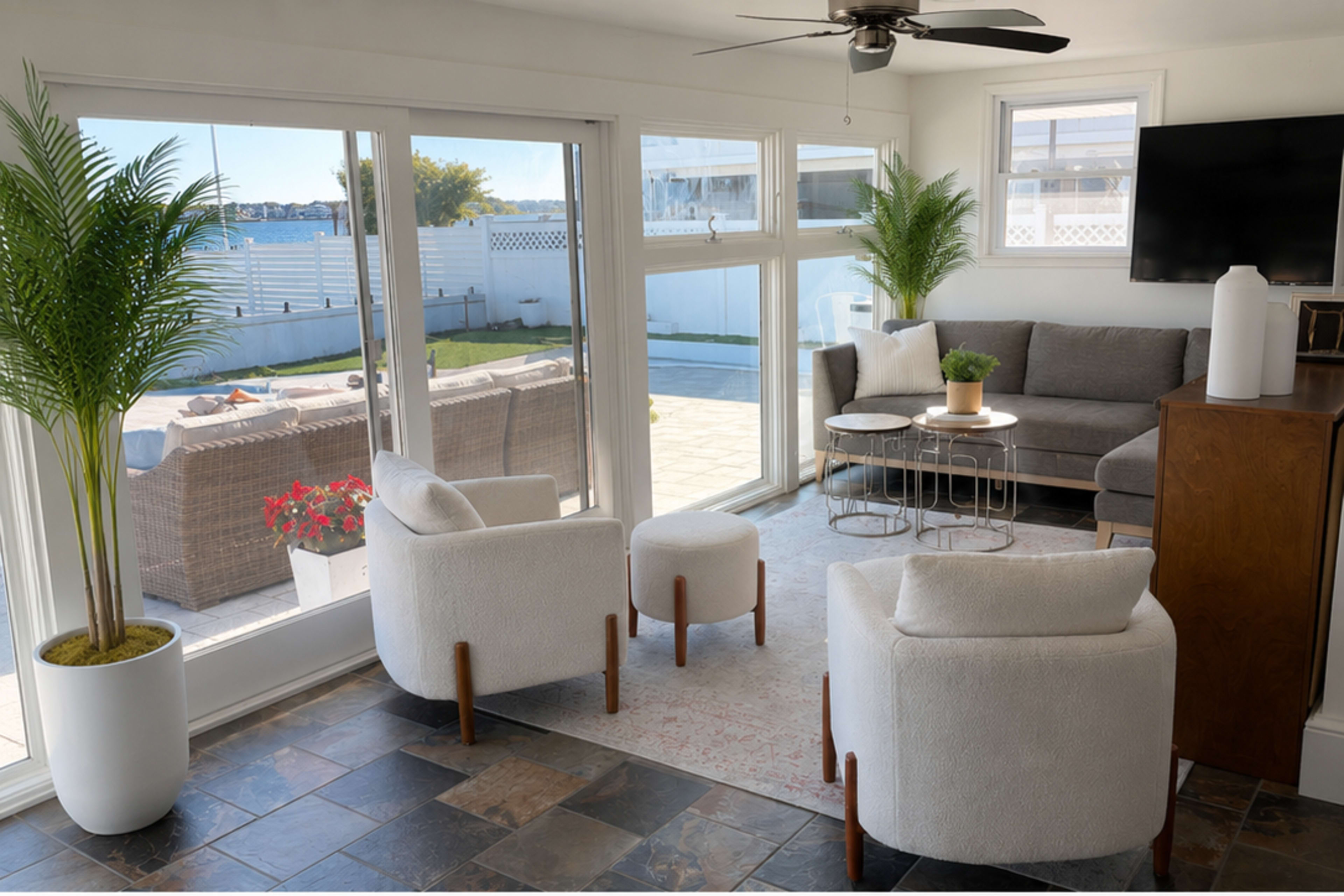 The image depicts a bright living room with large windows, modern furniture, a round coffee table, and a view of an outdoor patio area.