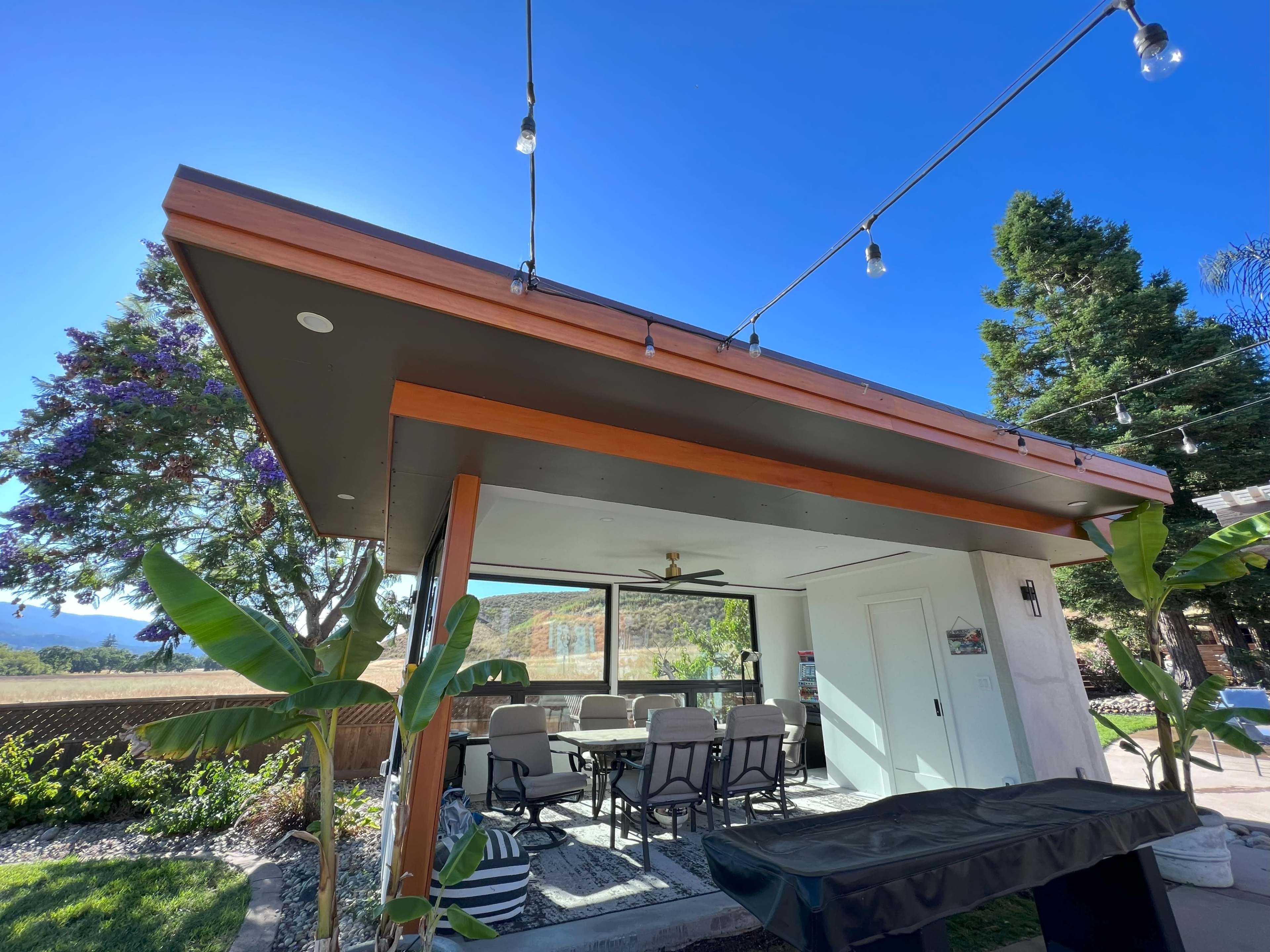 A modern outdoor patio with a dining area, surrounded by green plants and under a clear blue sky.