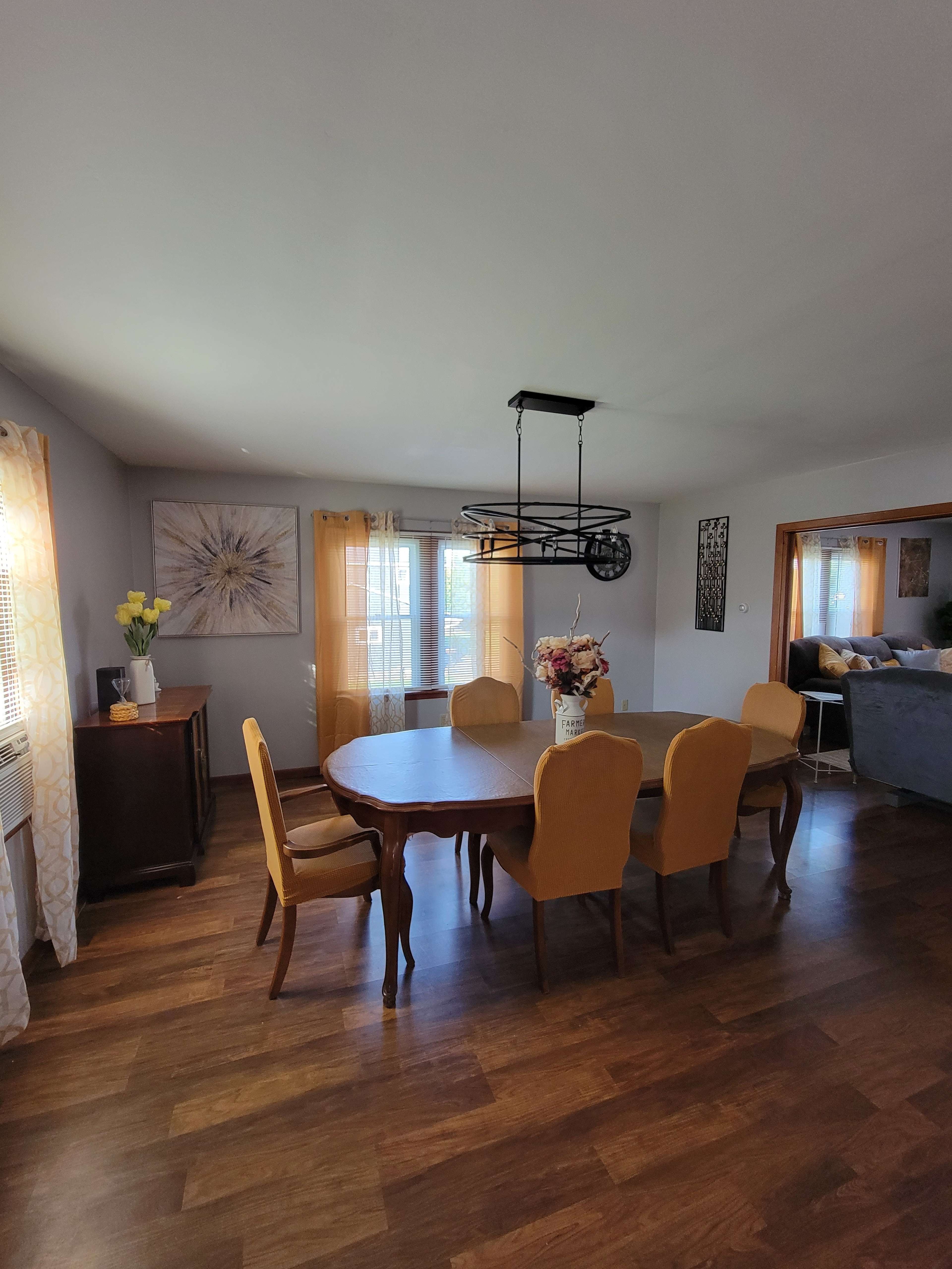 A dining area with a round wooden table surrounded by yellow upholstered chairs, featuring a floral centerpiece and large windows with sheer curtains.