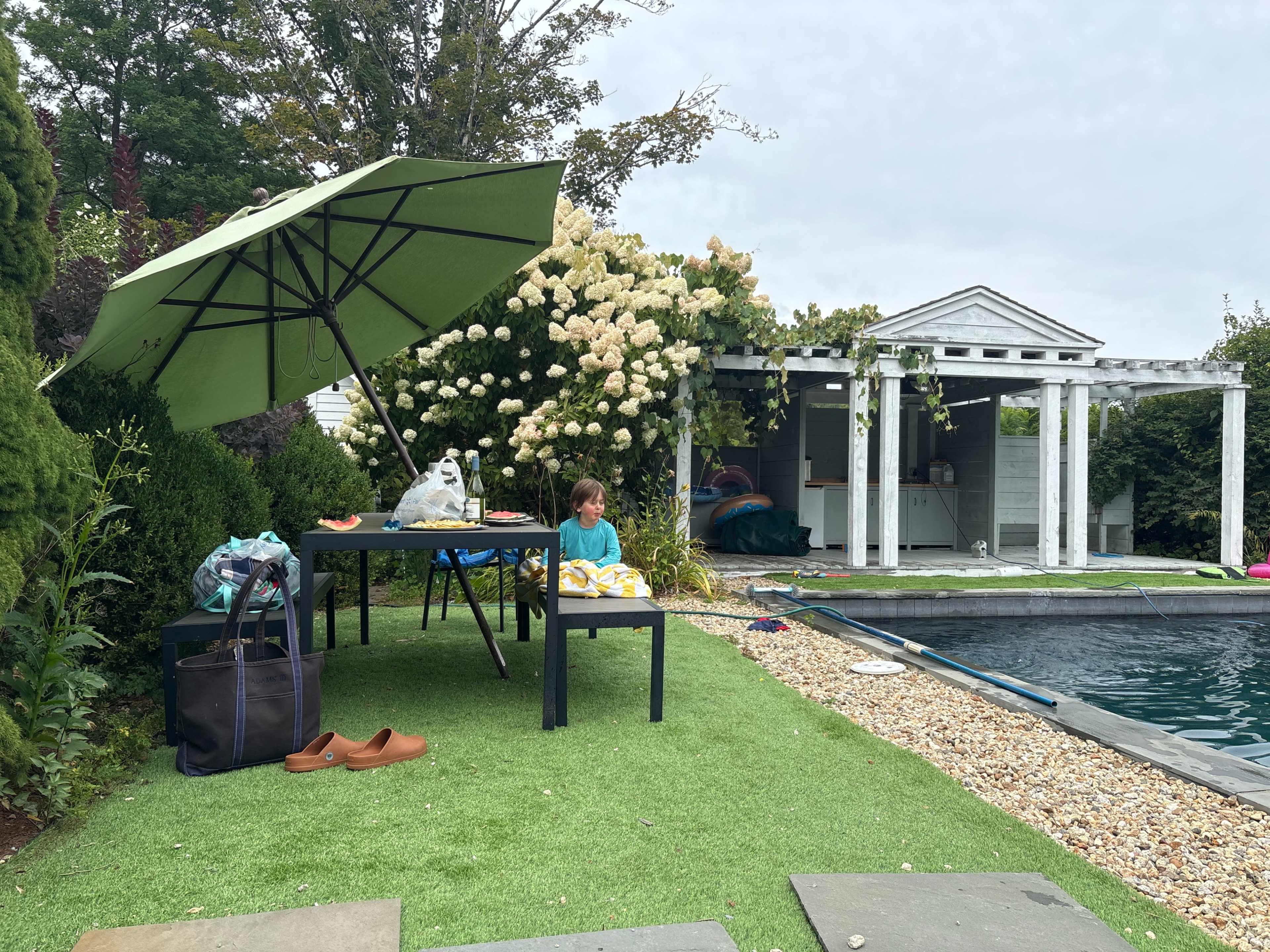 A child sits at a table under a large green umbrella next to a pool, with a white gazebo and flowering bushes in the background.