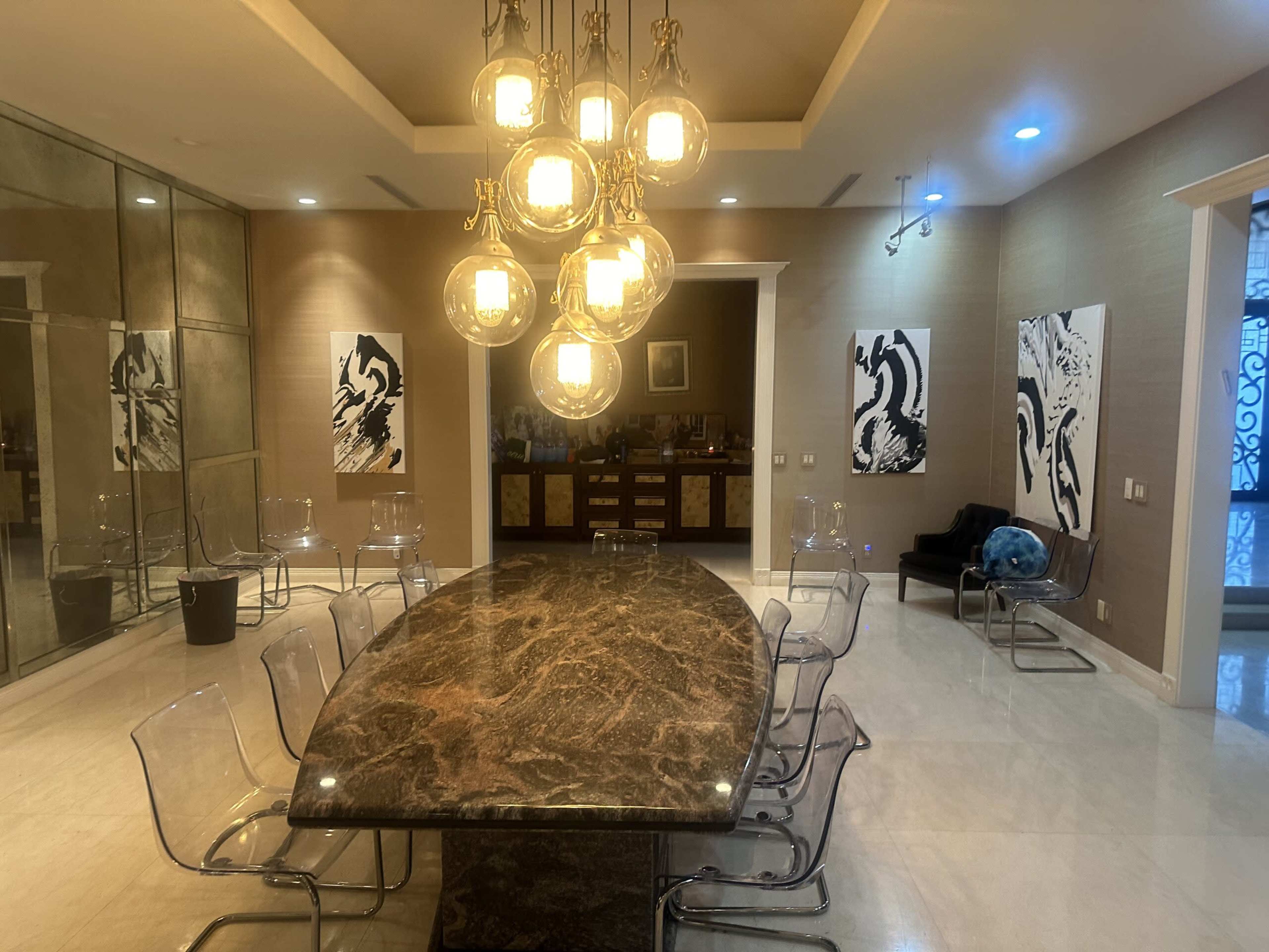 A spacious dining area features a large marble table surrounded by clear acrylic chairs, illuminated by a cluster of pendant lights, with abstract art hanging on the walls.