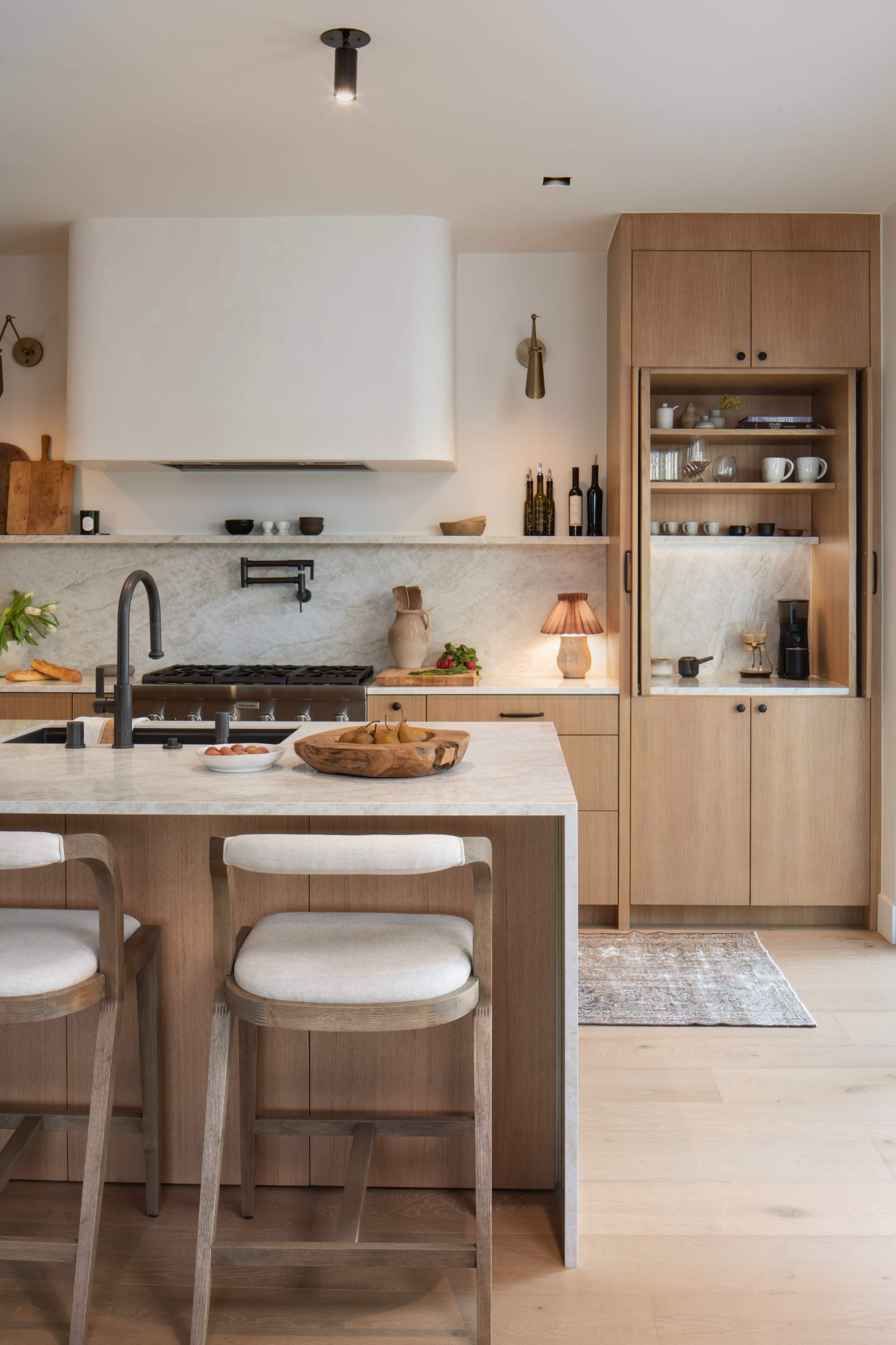 The image shows a modern kitchen with wooden cabinetry, a marble countertop, and two bar stools.
