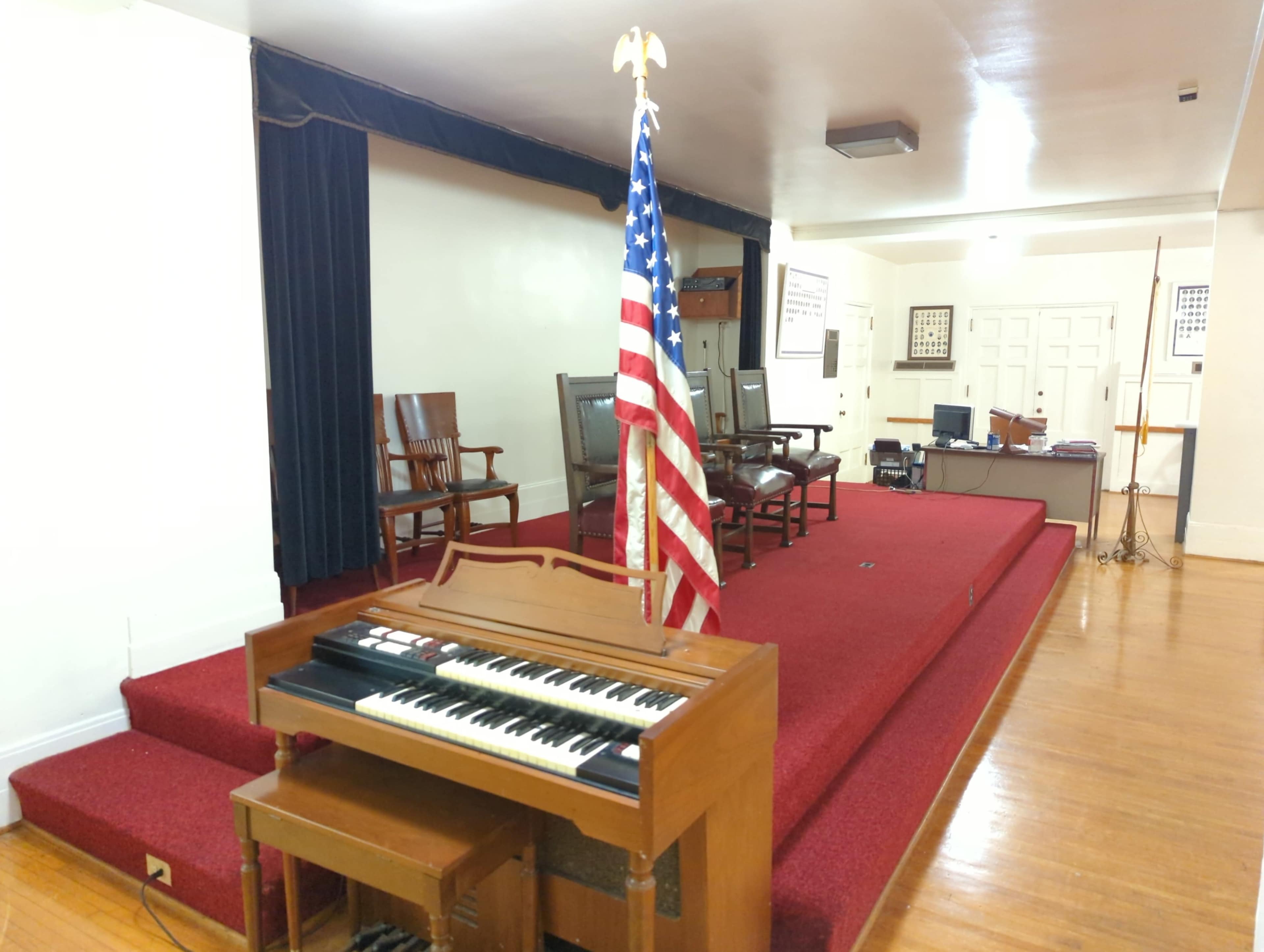 A wooden organ sits in front of a raised platform with several empty chairs and an American flag in a spacious meeting room.