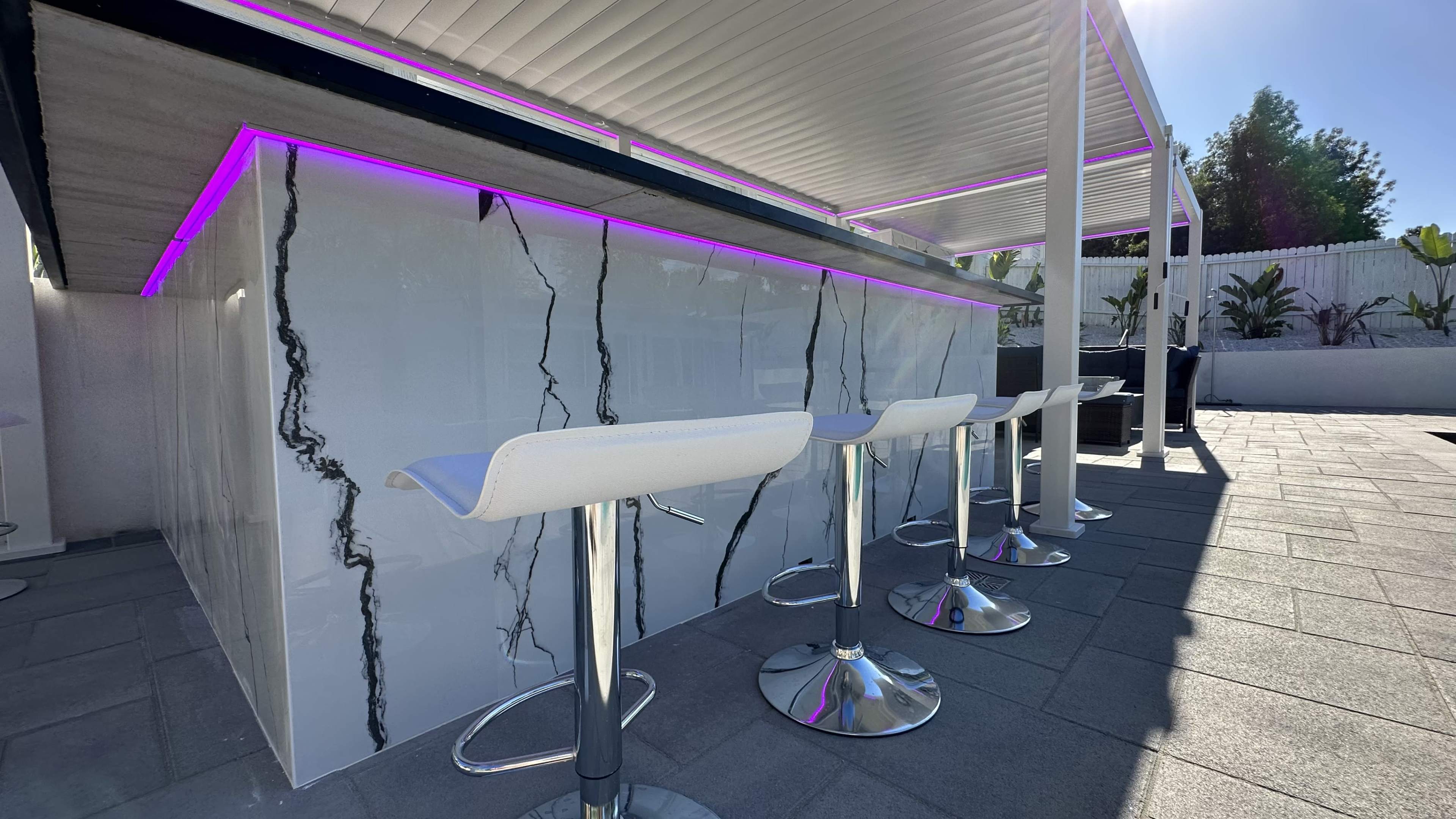 A modern outdoor bar with a marble-like design and backlit purple lighting, accompanied by white bar stools.