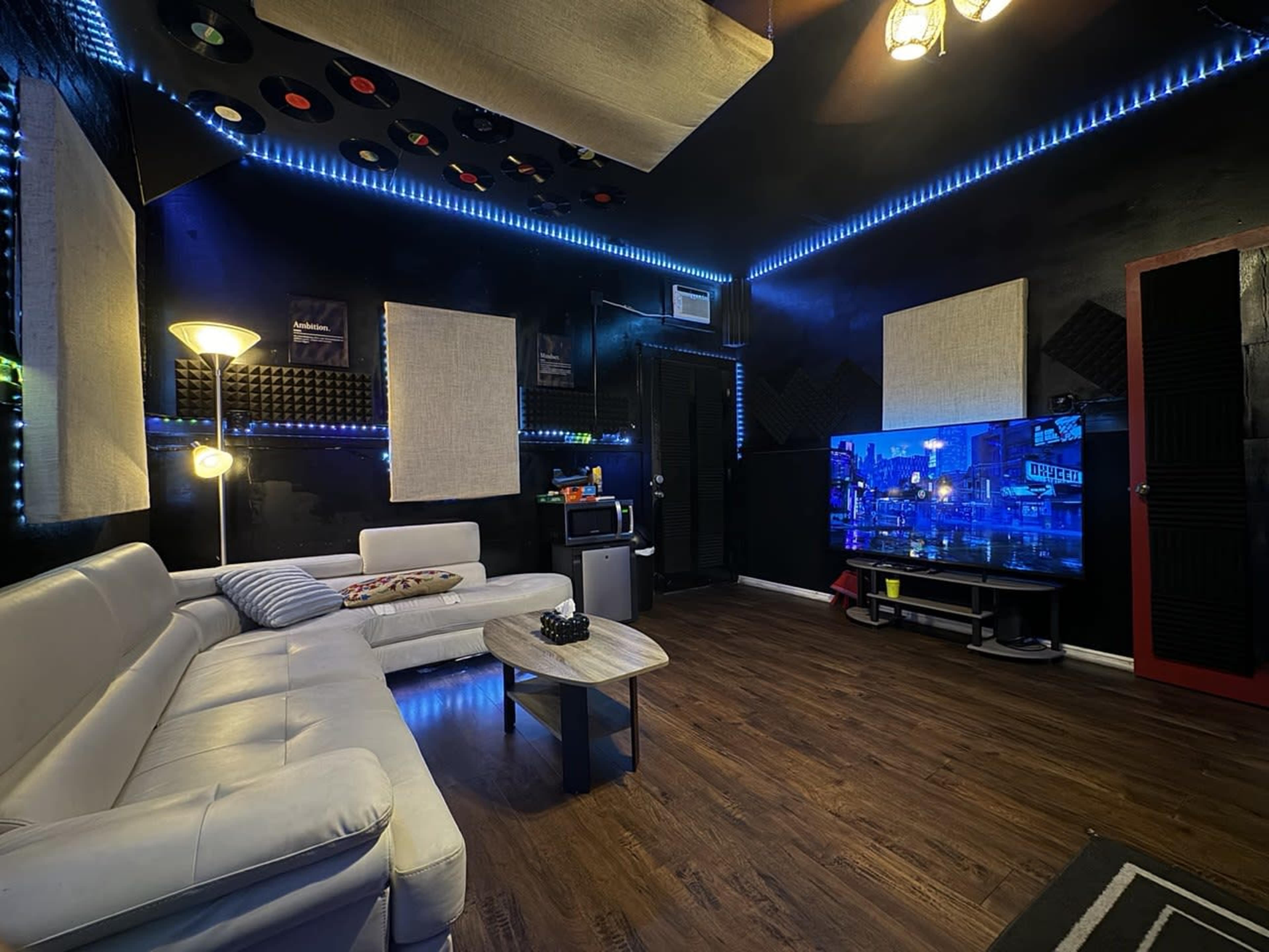 The image shows a music-themed lounge with a sectional sofa, a large flat-screen TV, and ambient lighting, featuring wall decor of vinyl records and soundproof panels.