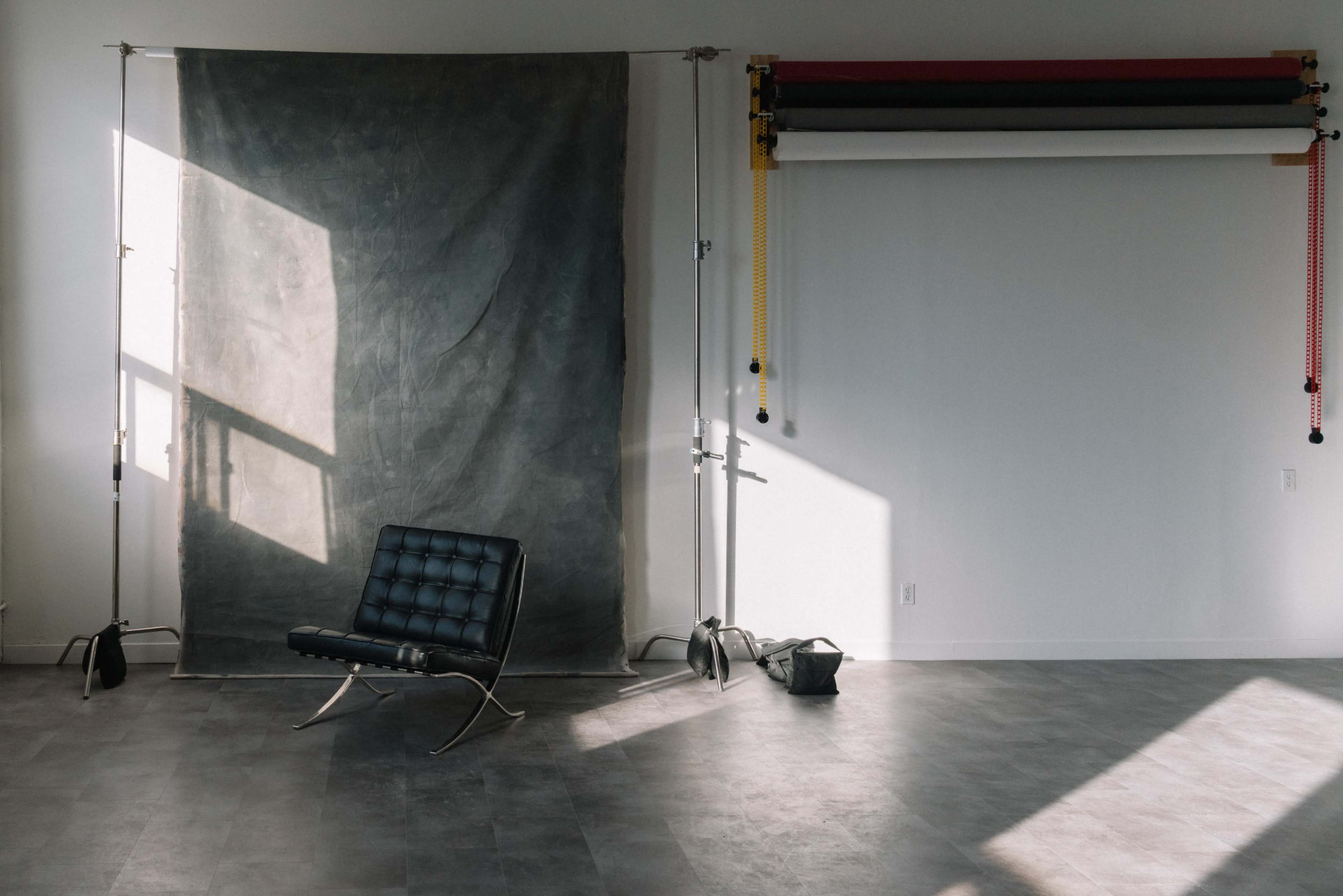 A black chair is positioned in front of a gray backdrop, with sunlight casting a shadow on the floor in a spacious room.