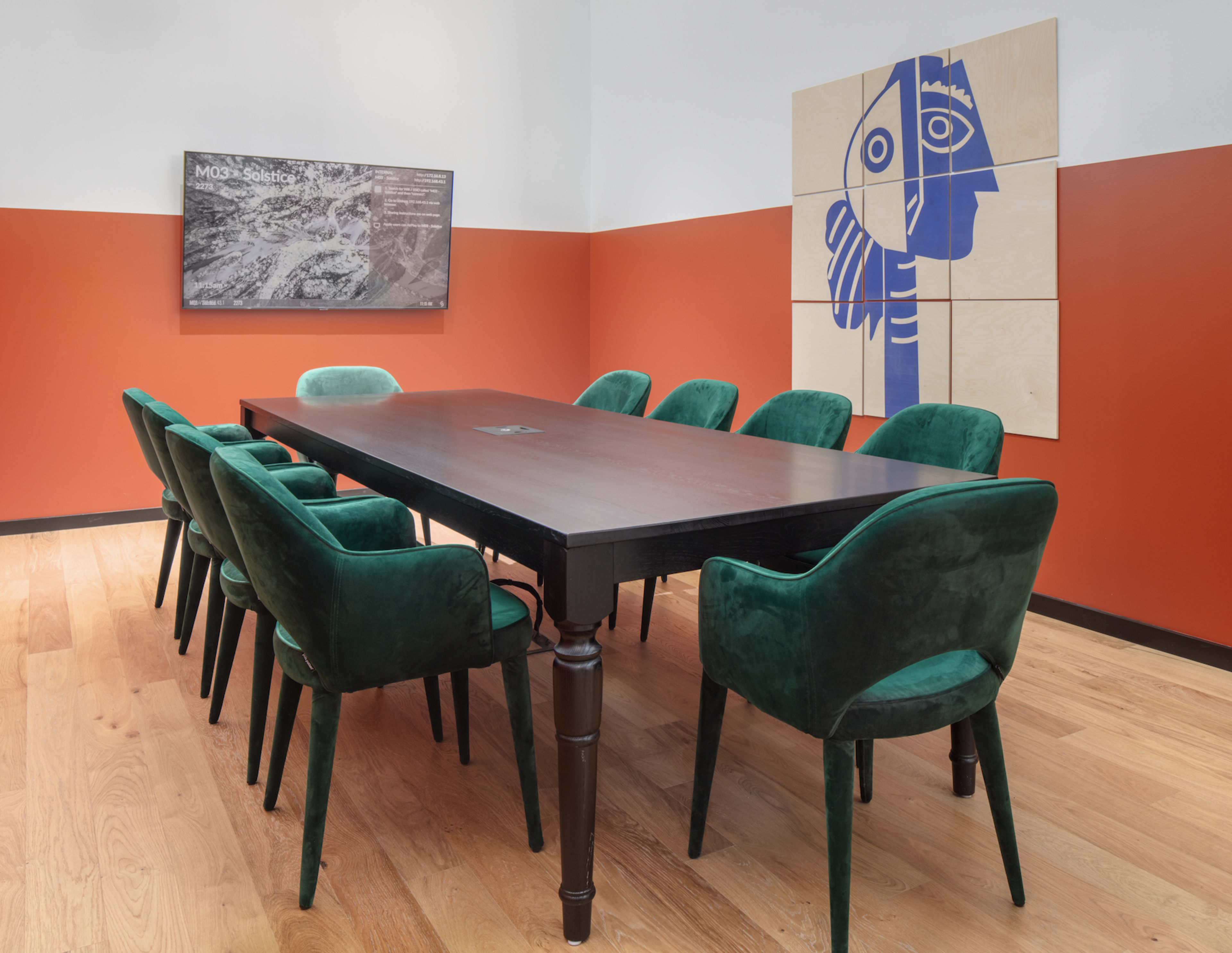 A long wooden conference table is surrounded by green upholstered chairs in a room with orange and white walls, featuring a large abstract artwork and a screen displaying information on the wall.