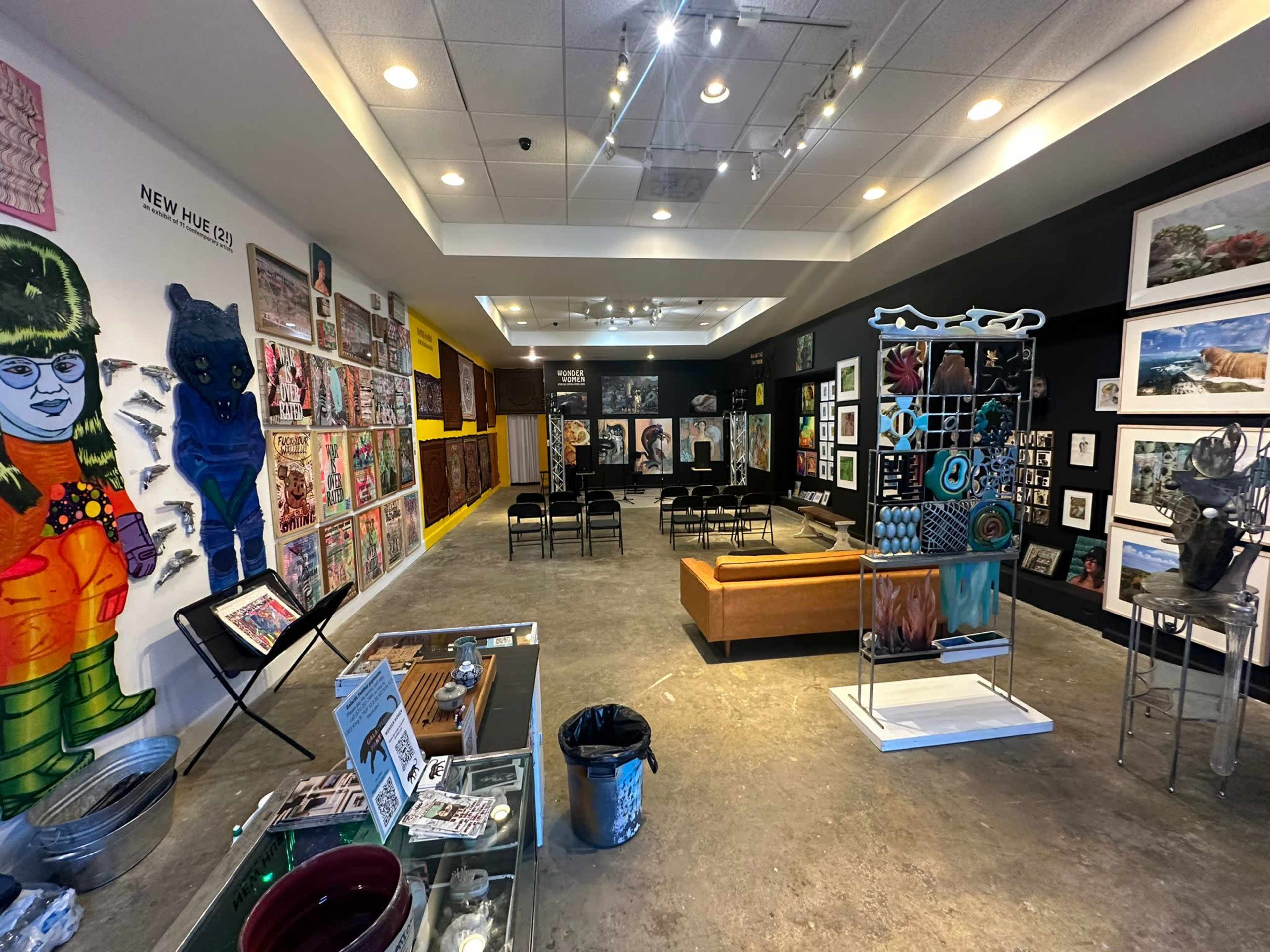 An art gallery featuring colorful wall displays of various artworks and sculptures, with chairs arranged for a viewing area.
