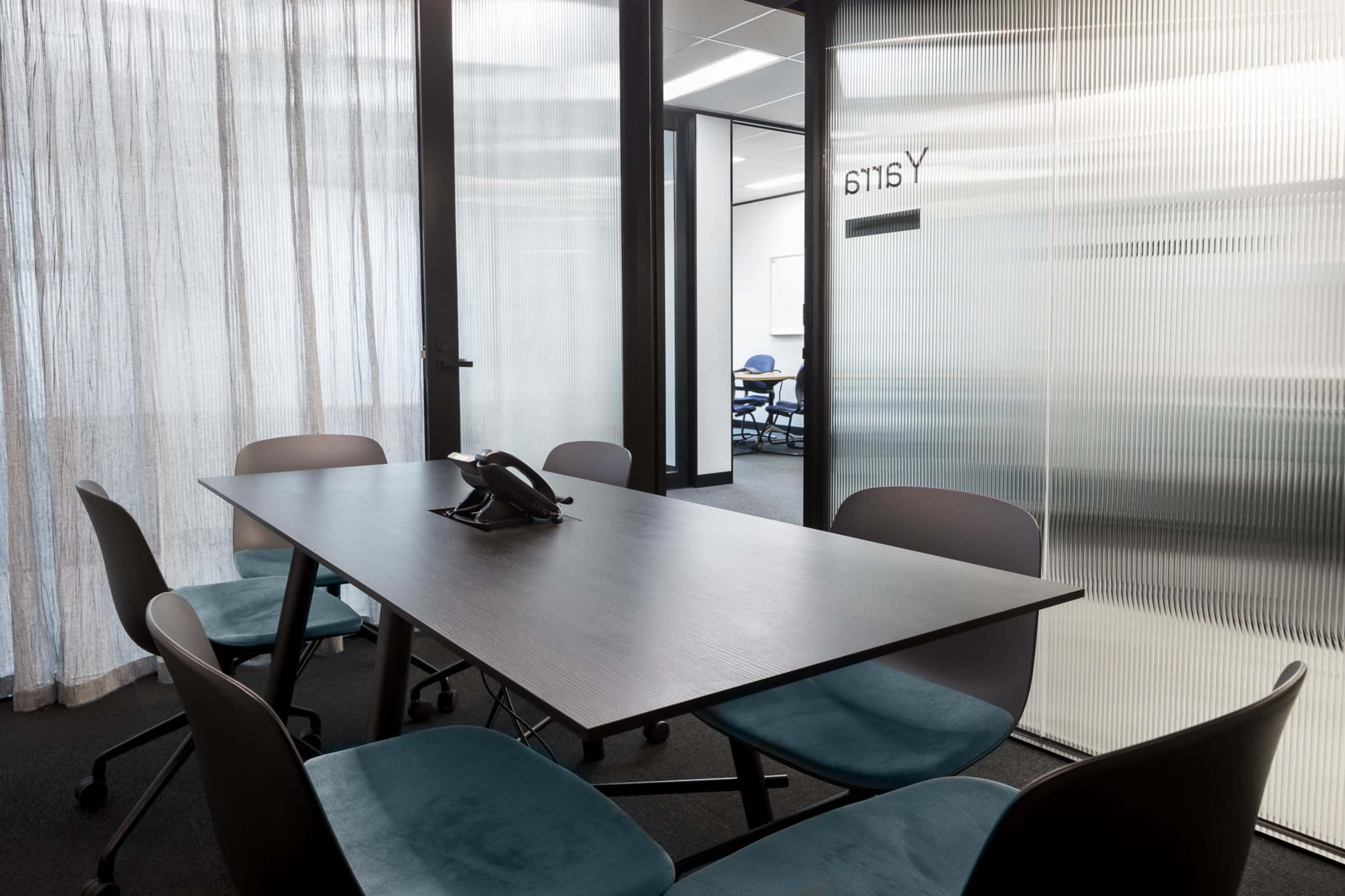 A modern conference room features a rectangular table with a telephone and six teal chairs, surrounded by glass sliding doors with frosted panels.
