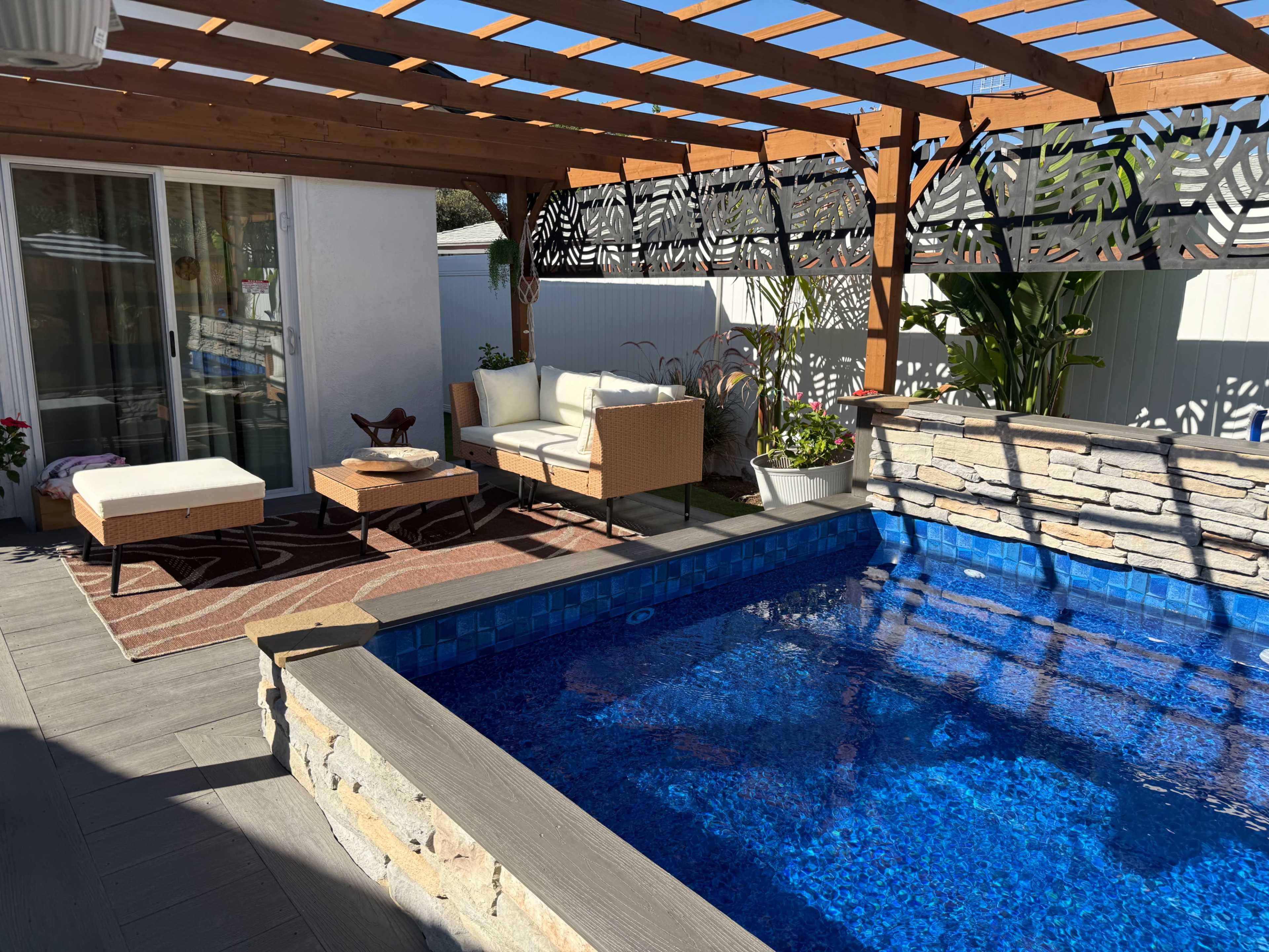 The image shows a small outdoor space featuring a pool surrounded by a deck, with seating arrangements under a wooden pergola.