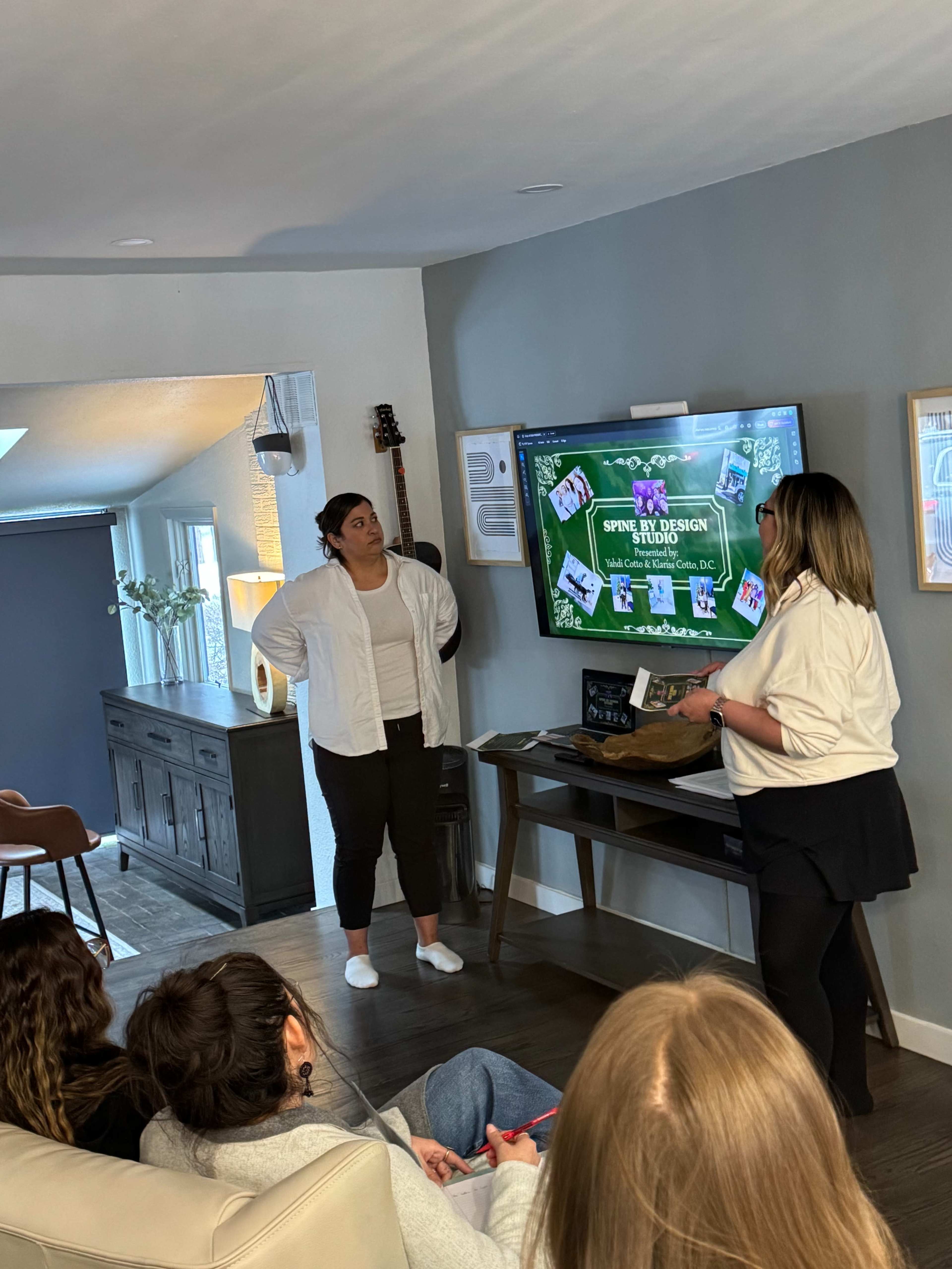 Two women stand in front of a television displaying a presentation, while a small group sits on a couch observing in a modern living room setting.