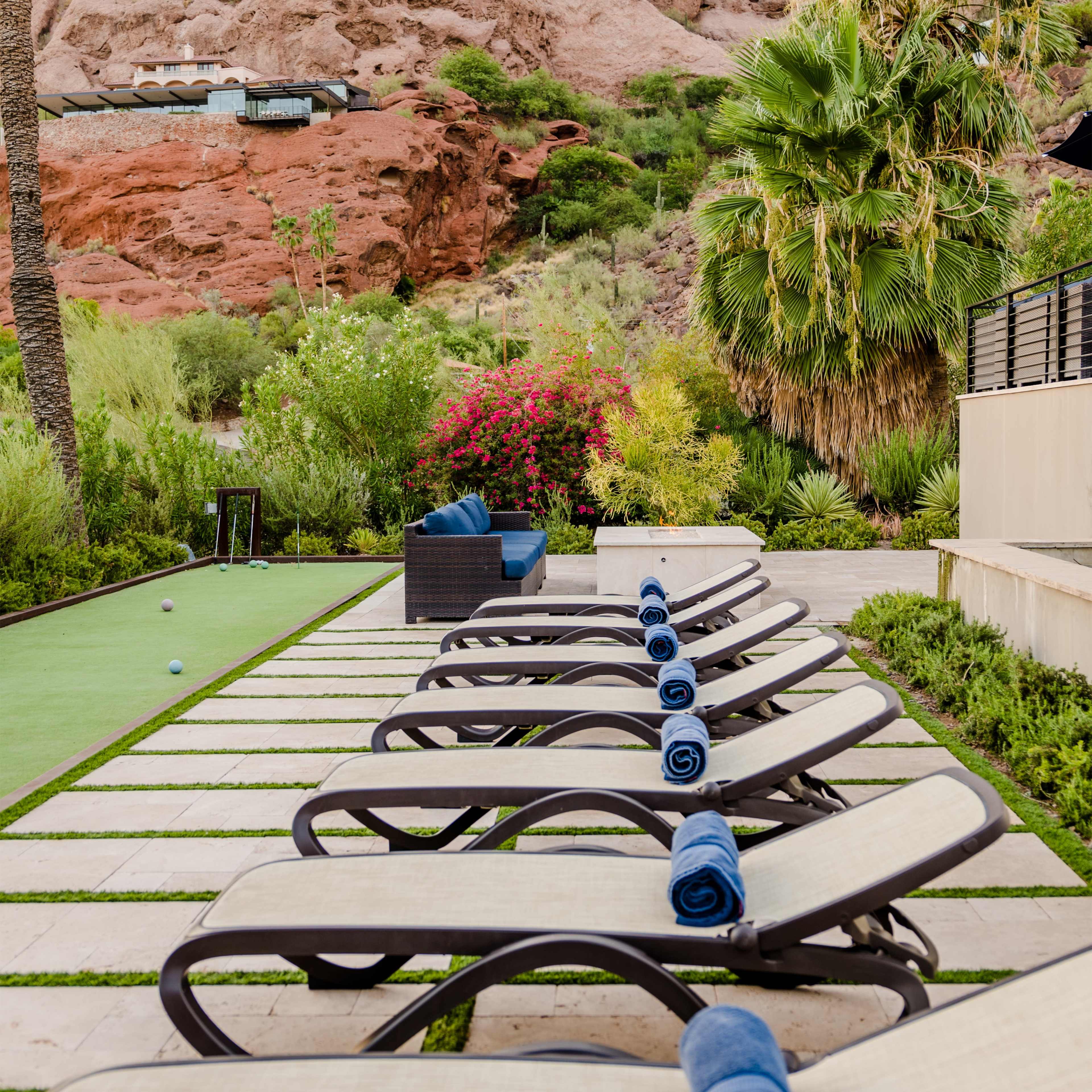 A row of lounge chairs with blue towels placed on them, beside a well-maintained green lawn and surrounded by tropical plants and rocky terrain.