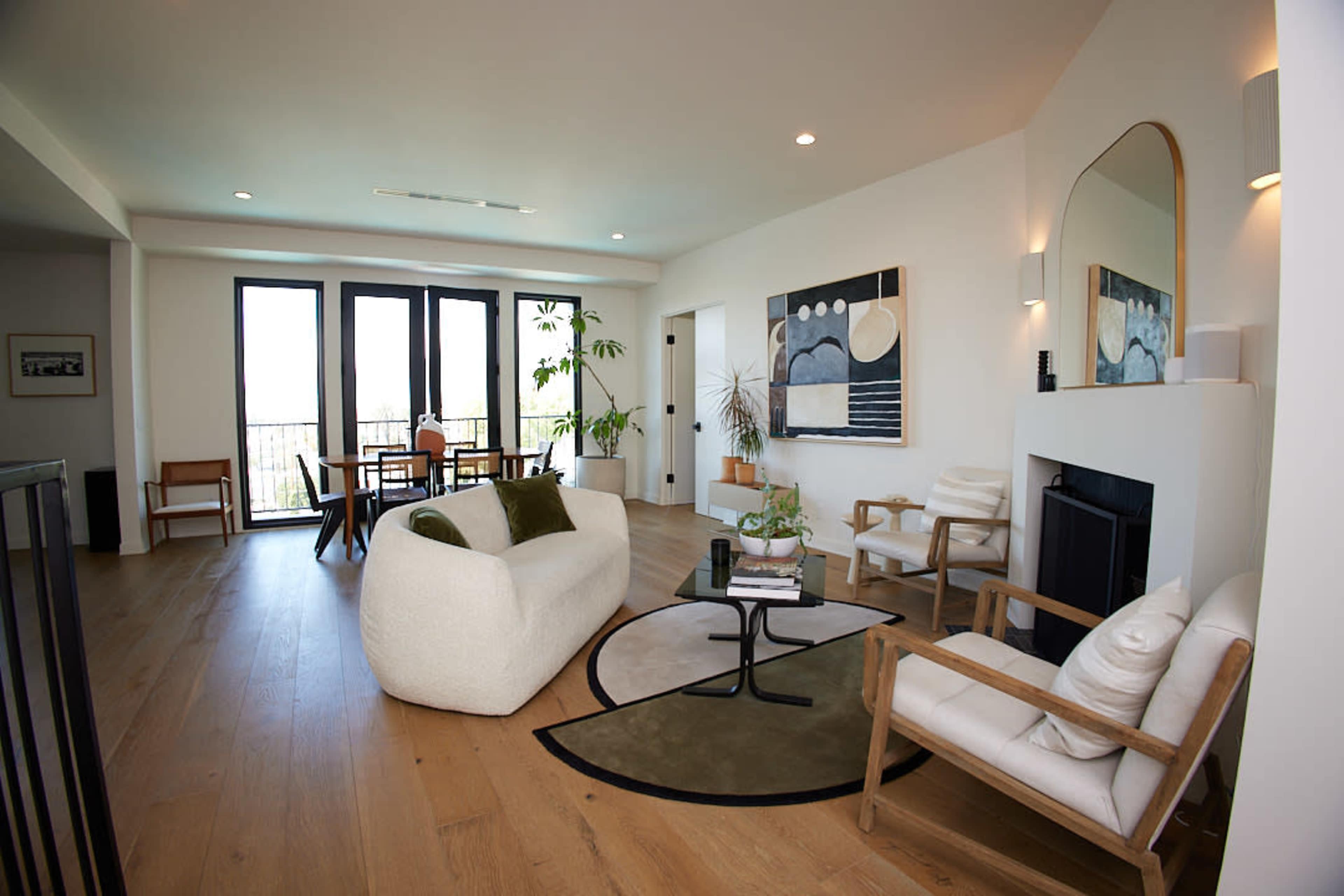 The spacious living room features a cozy white sofa, two armchairs, a round coffee table, and large windows that provide natural light.