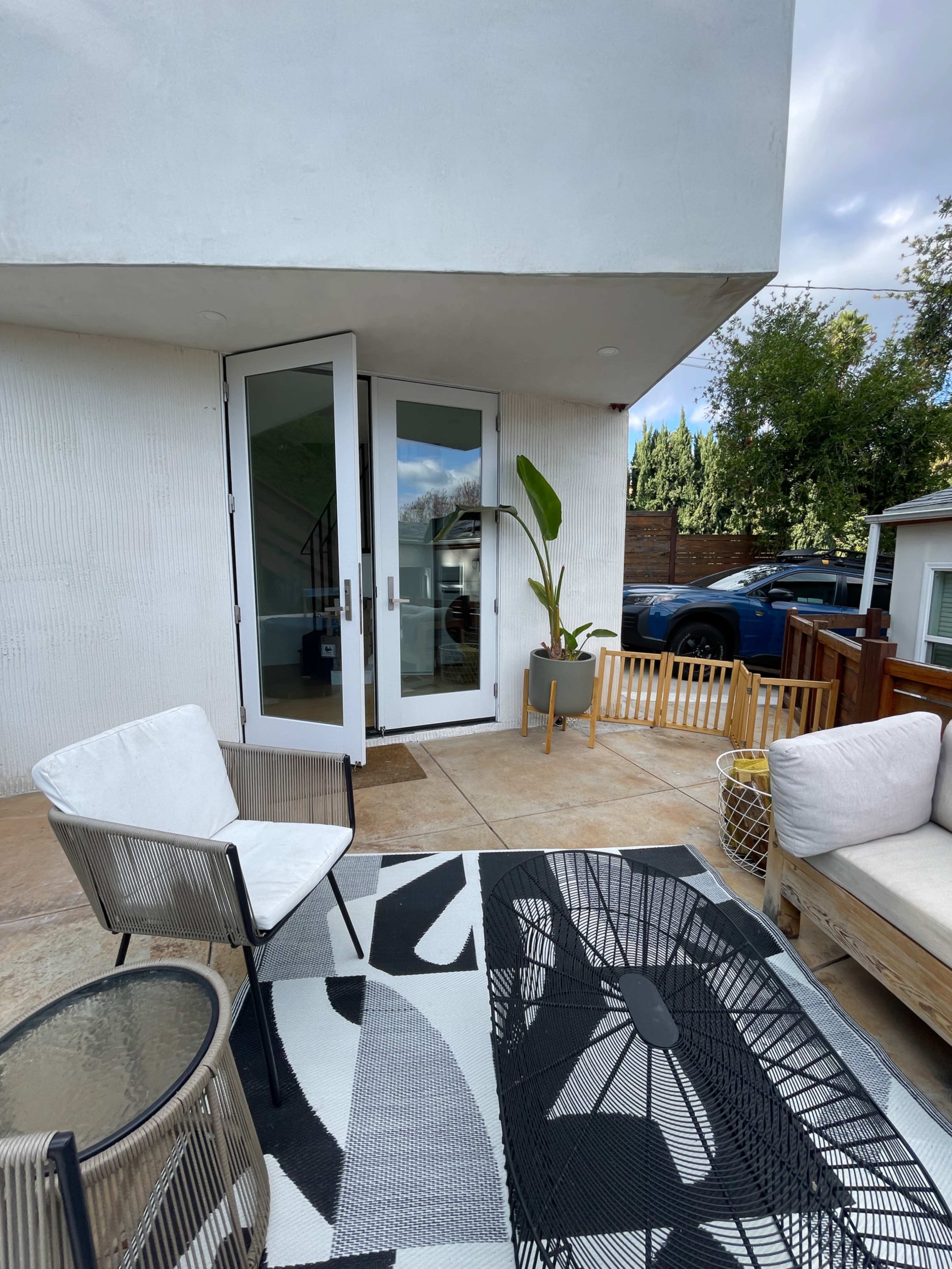 The image shows a small outdoor seating area with modern furniture, including two chairs and a couch, next to a glass door leading into a building.