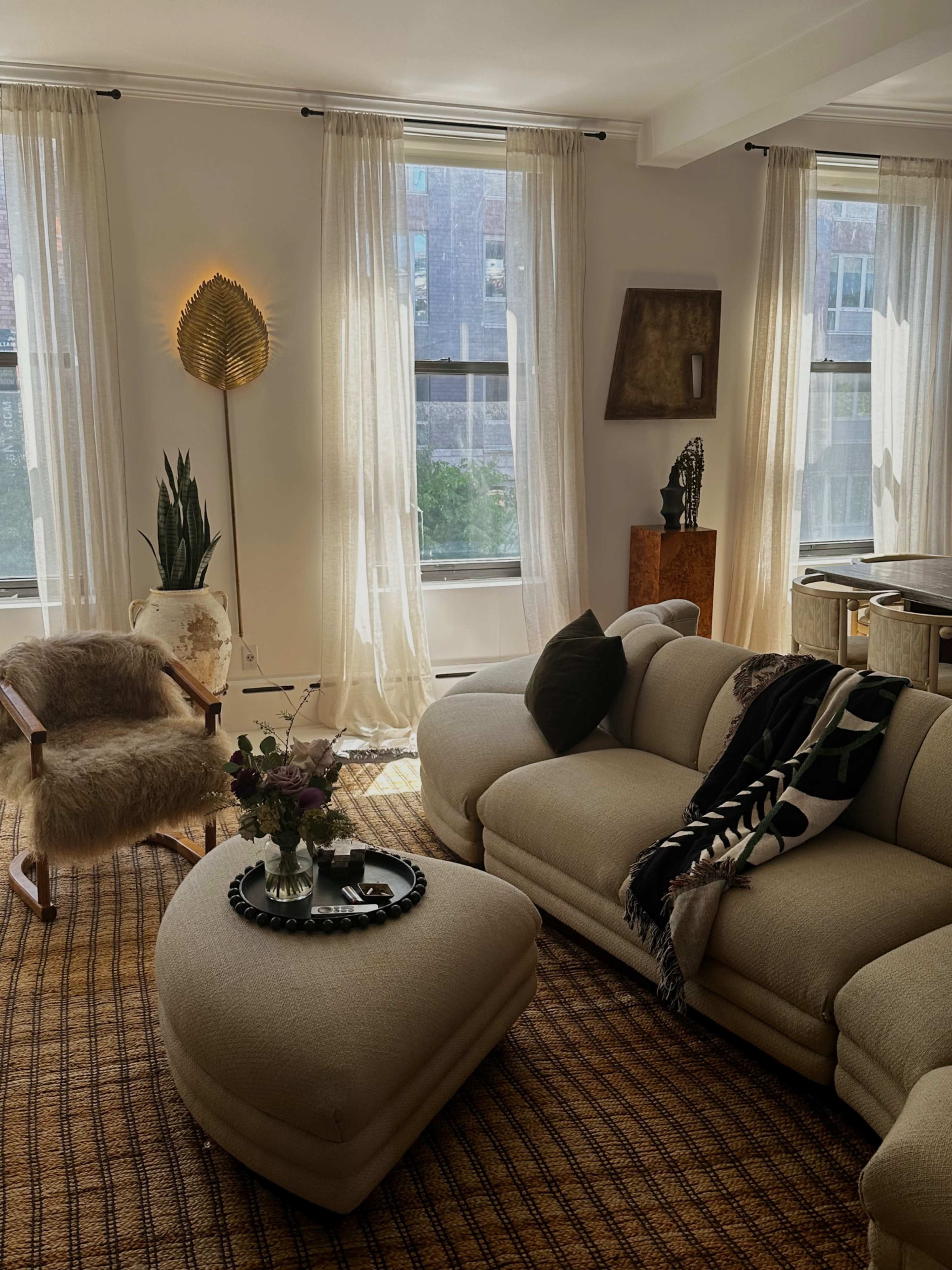 A living room features a light beige sectional sofa, a round coffee table with a vase of flowers, and large windows draped with sheer curtains.