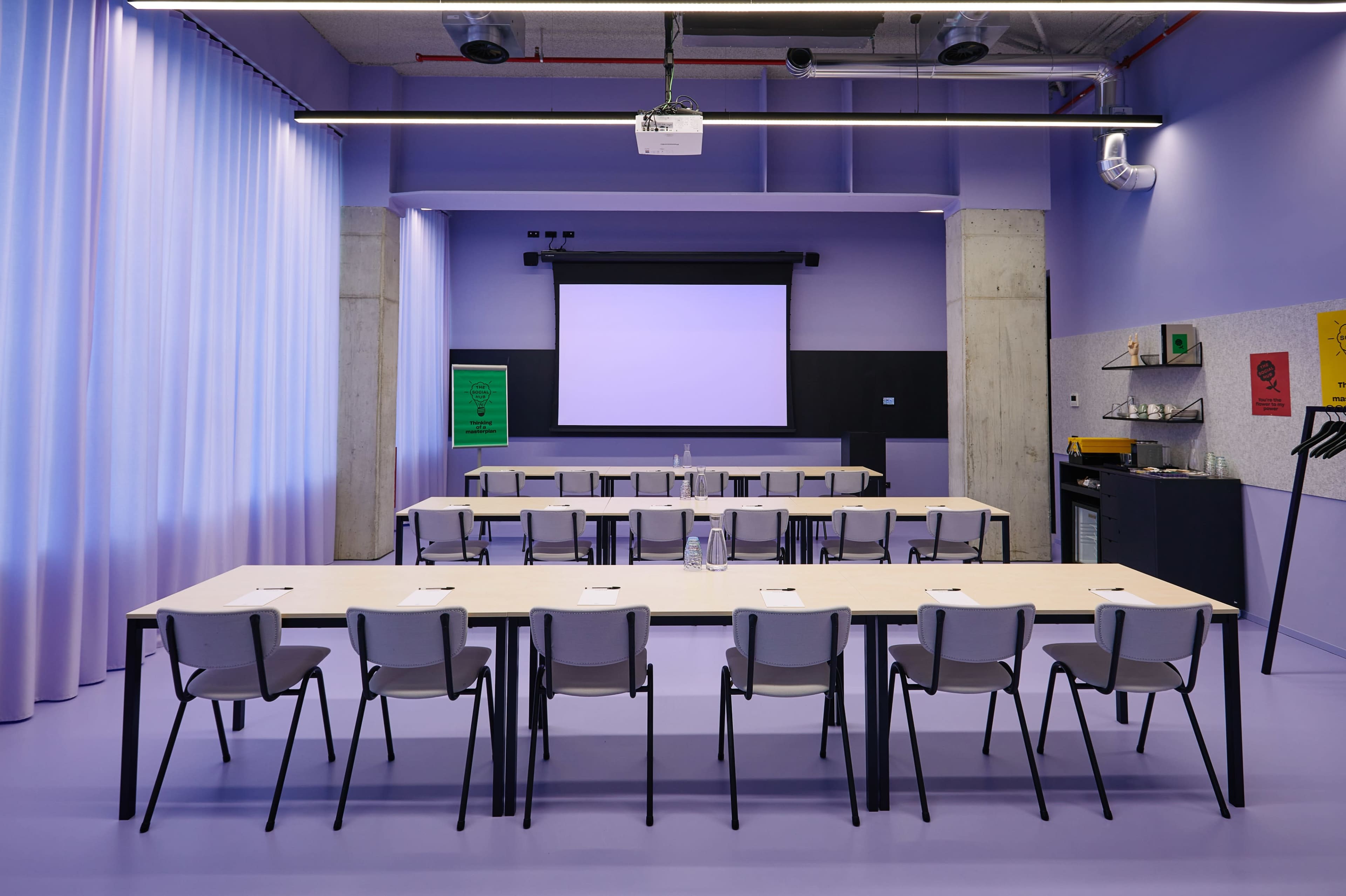 A modern conference room features a long table with chairs arranged in front of a projection screen on a pastel-colored wall.