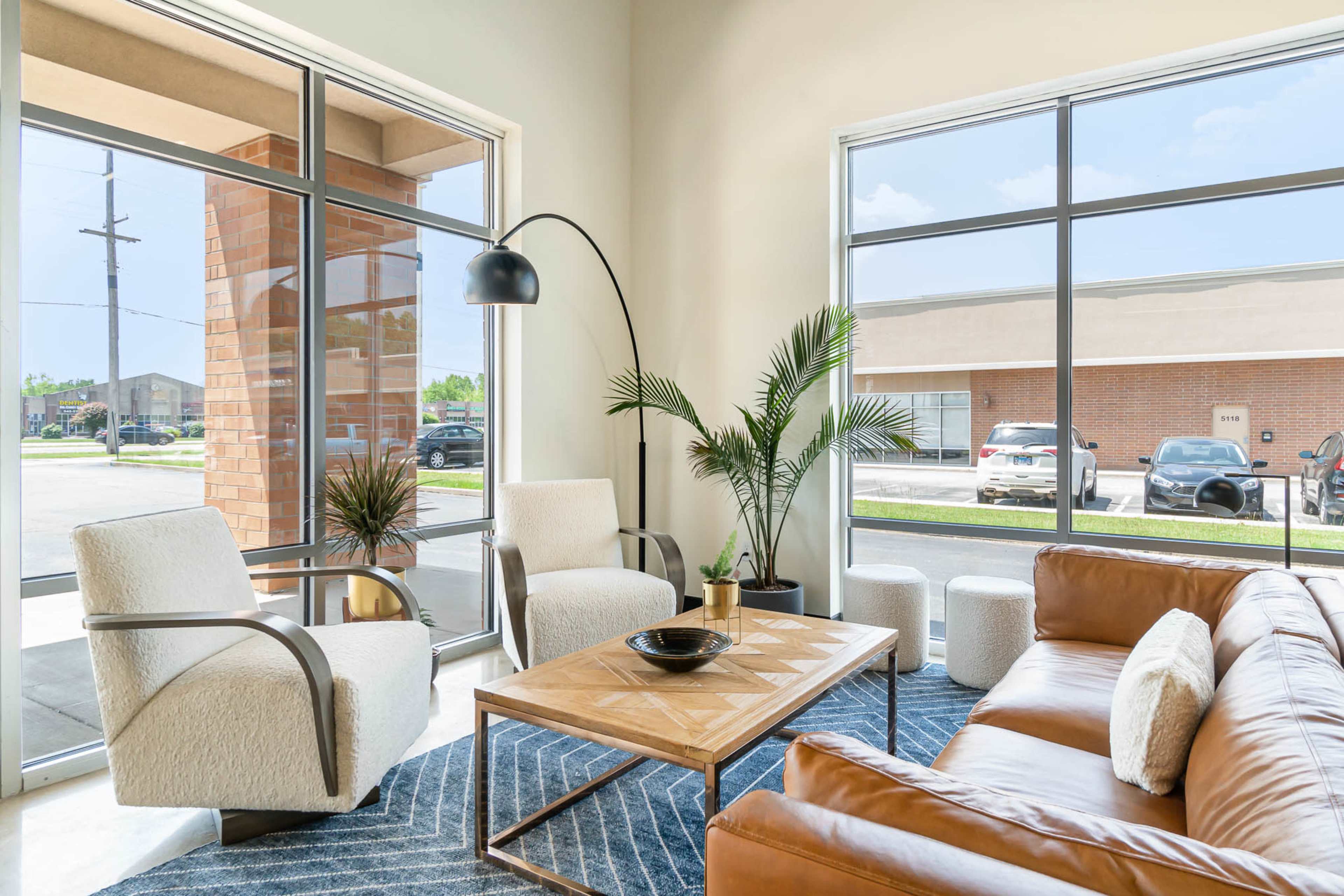 A modern seating area with a brown leather sofa, two armchairs, a coffee table, and large windows letting in natural light.