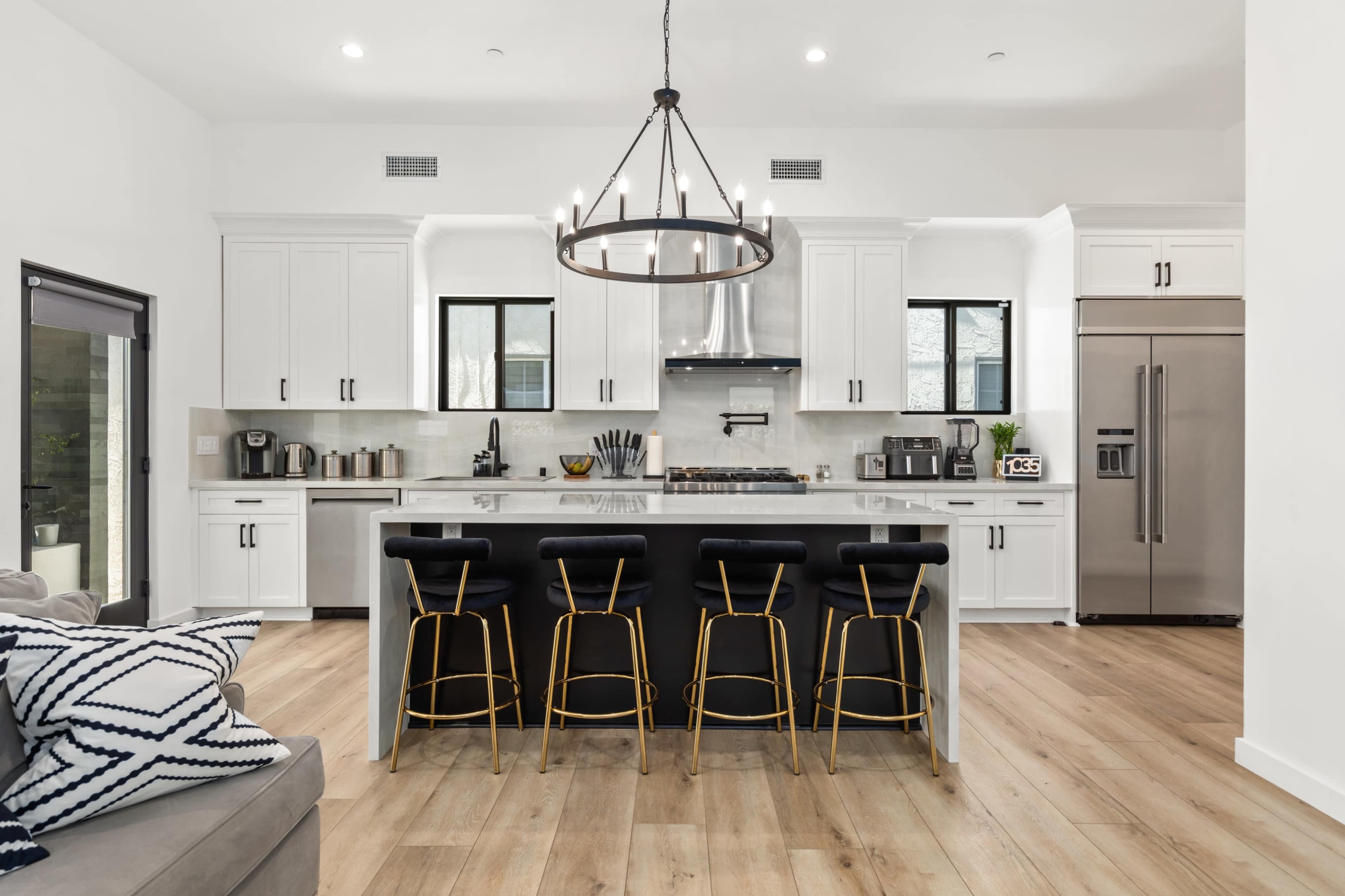 The image shows a modern kitchen with white cabinetry, stainless steel appliances, a large island with seating, and hardwood flooring.