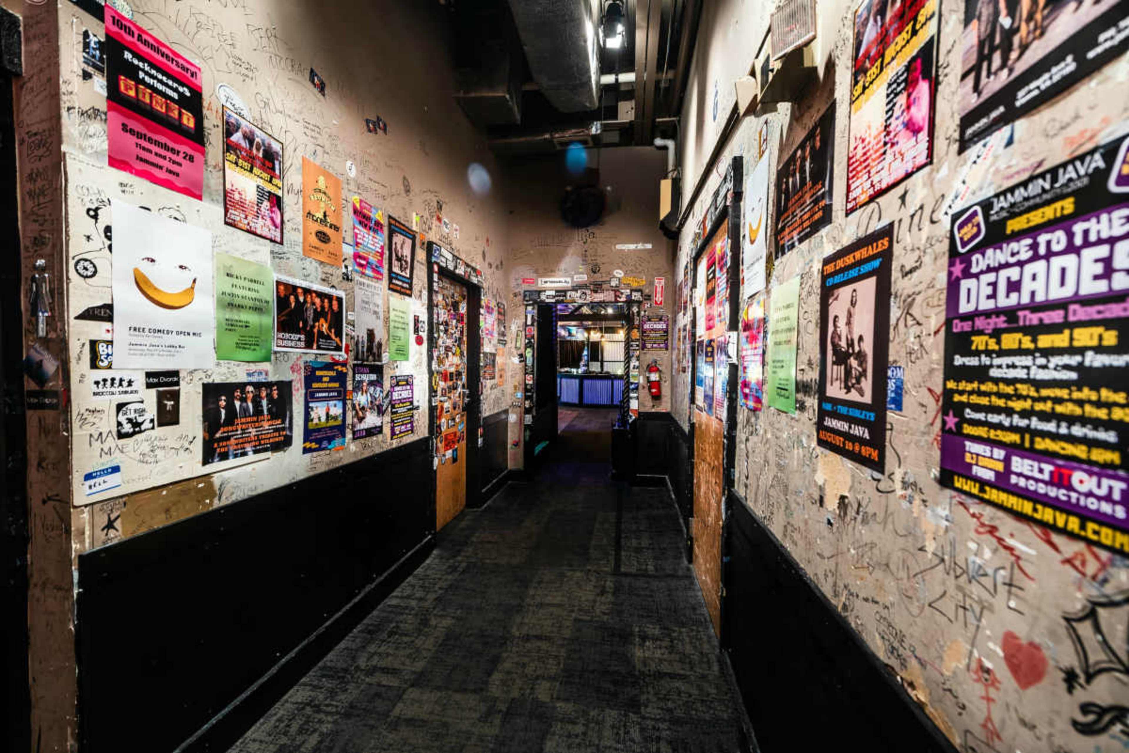The image shows a narrow hallway lined with posters and graffiti-covered walls leading to a door at the far end.