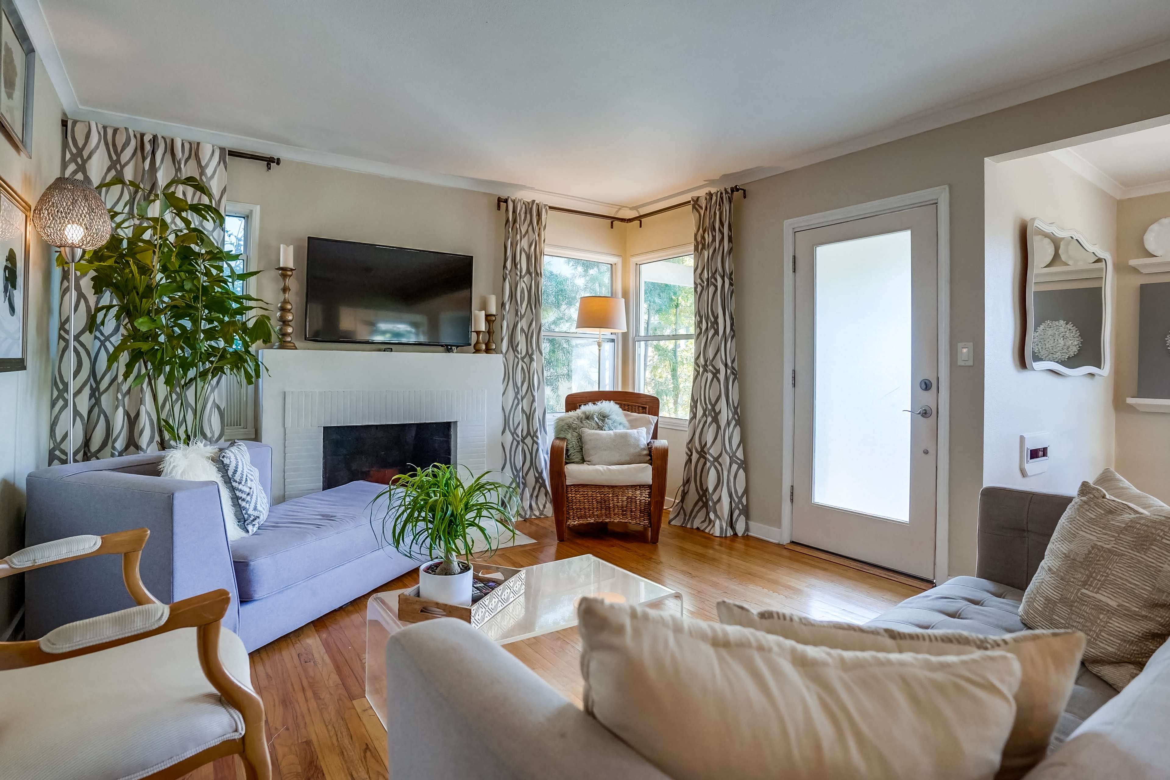 The image shows a cozy living room featuring a television mounted on the wall, a fireplace, large windows, and a mix of seating options, including a gray sofa and a wicker chair.