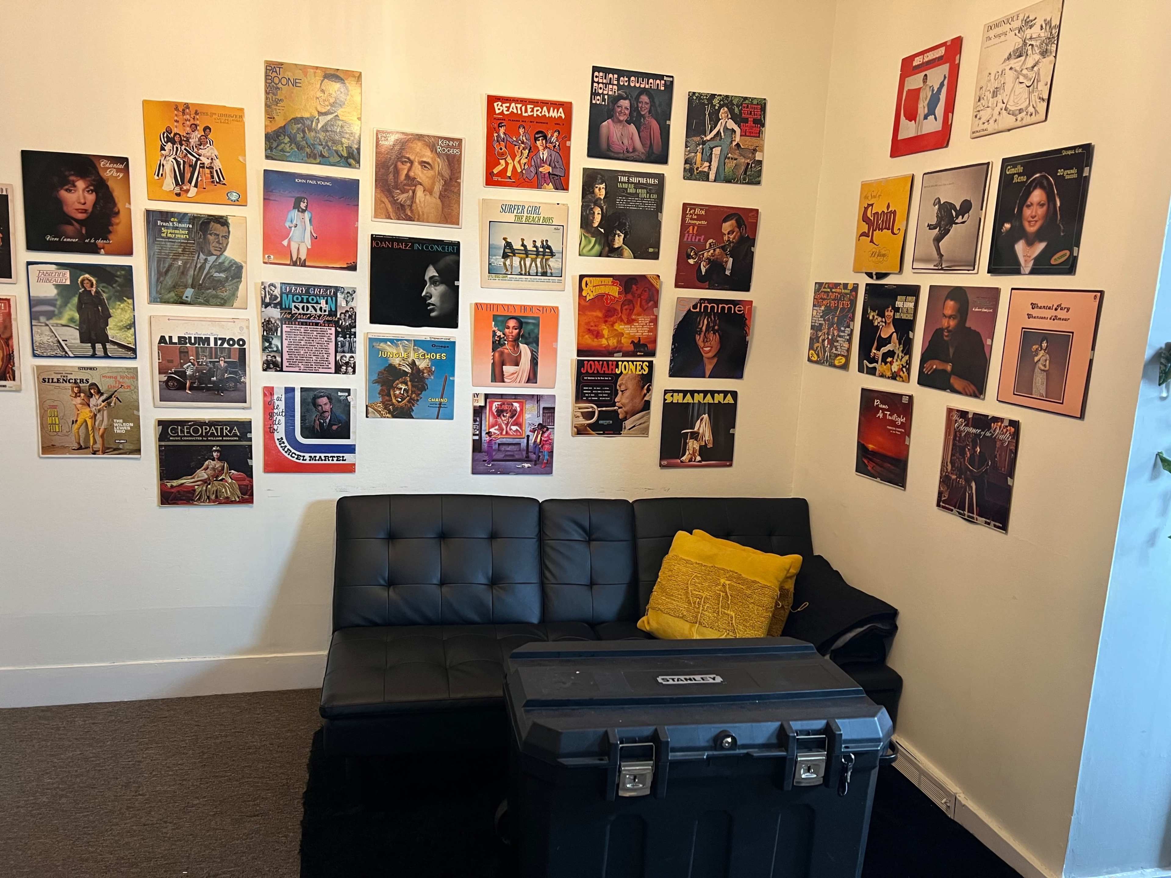 The image shows a cozy room with a black sofa and a black storage box, surrounded by various vintage vinyl record covers displayed on the walls.