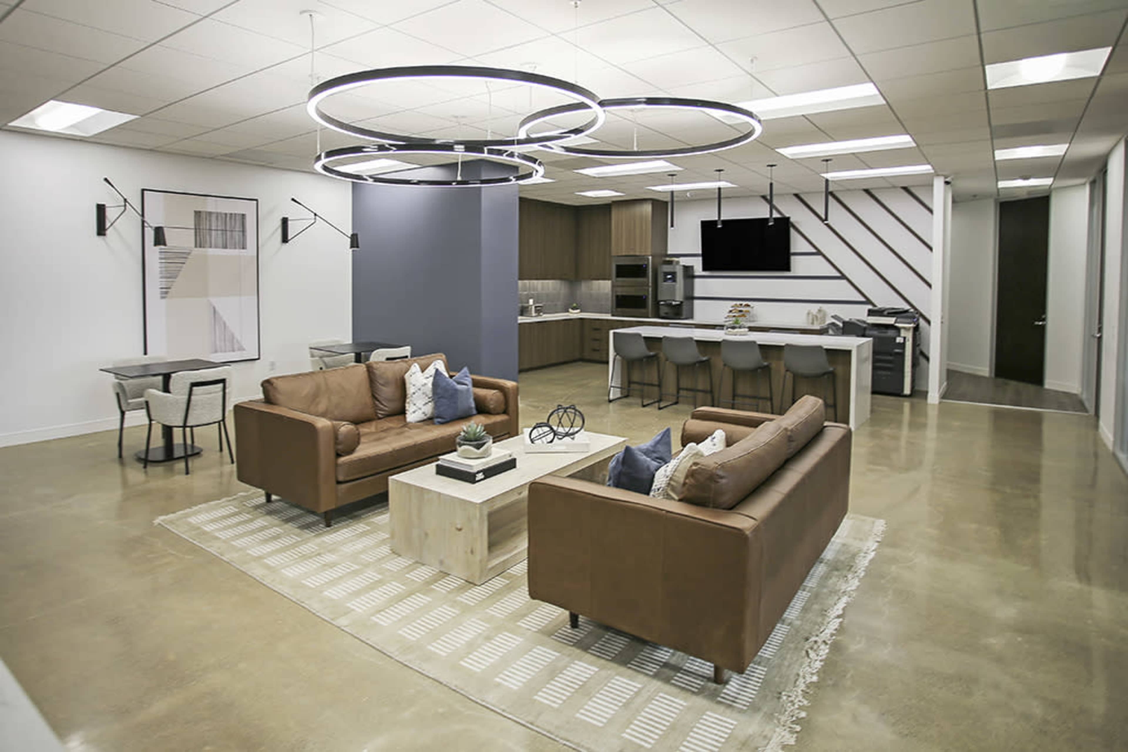 The image shows a modern lounge area with two brown sofas, a coffee table, and a dining table, adjacent to a kitchen featuring dark cabinets and a mounted television.