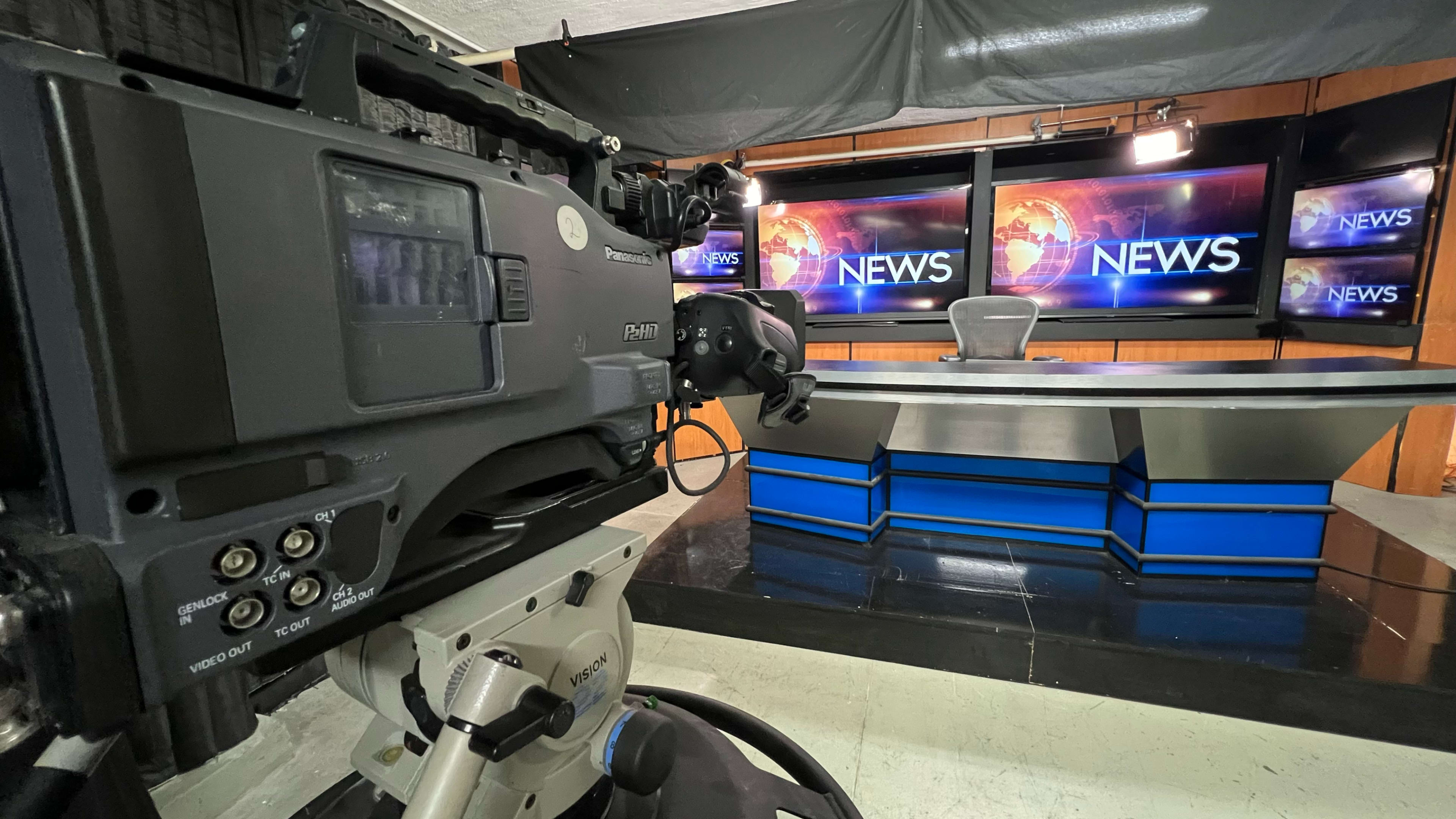 The image shows a television news studio with a camera in the foreground and a news desk with monitors displaying the word "NEWS" in the background.