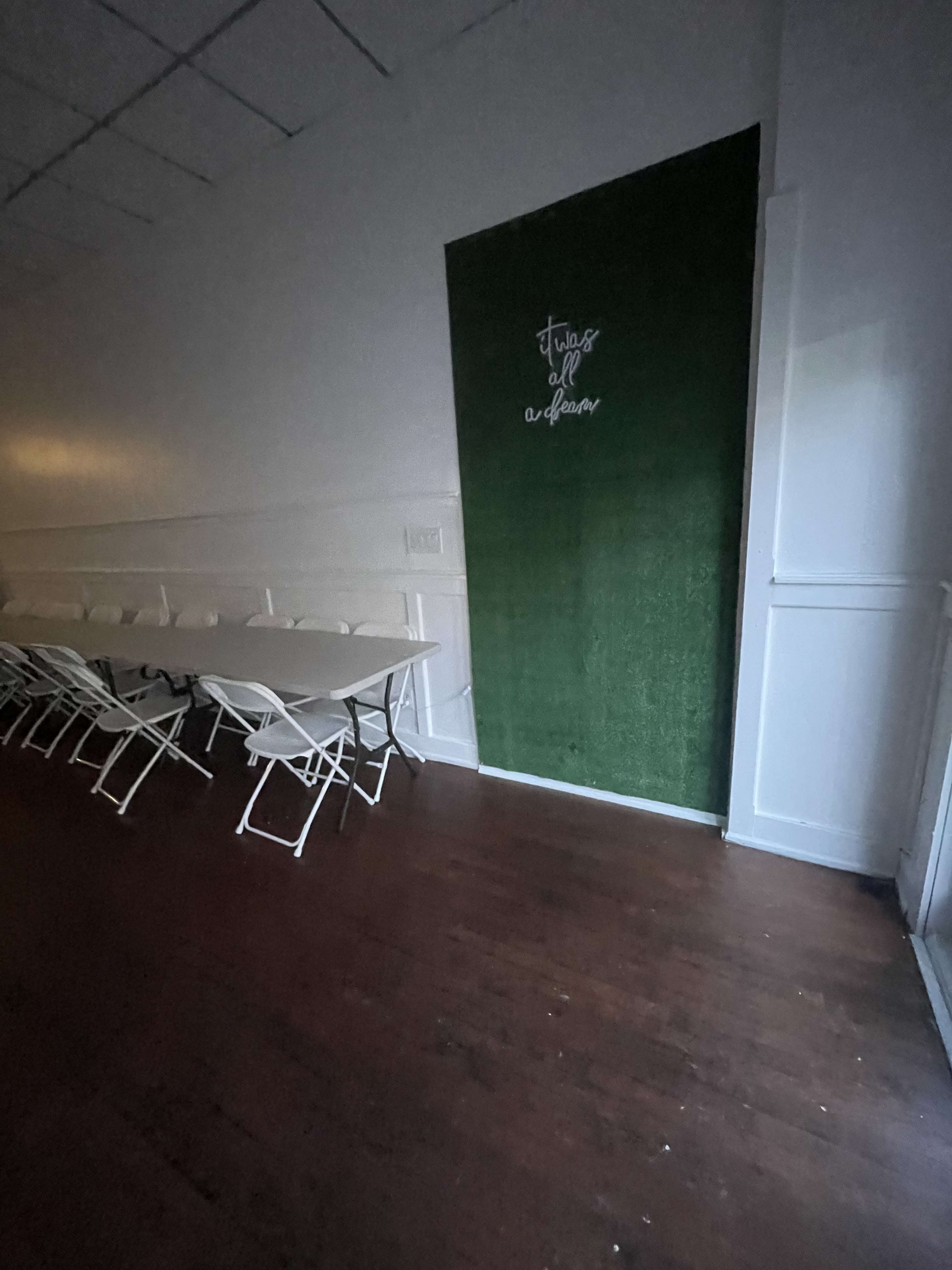 The image shows a dimly lit room with a long white table and folding chairs arranged around it, next to a green wall featuring white text.