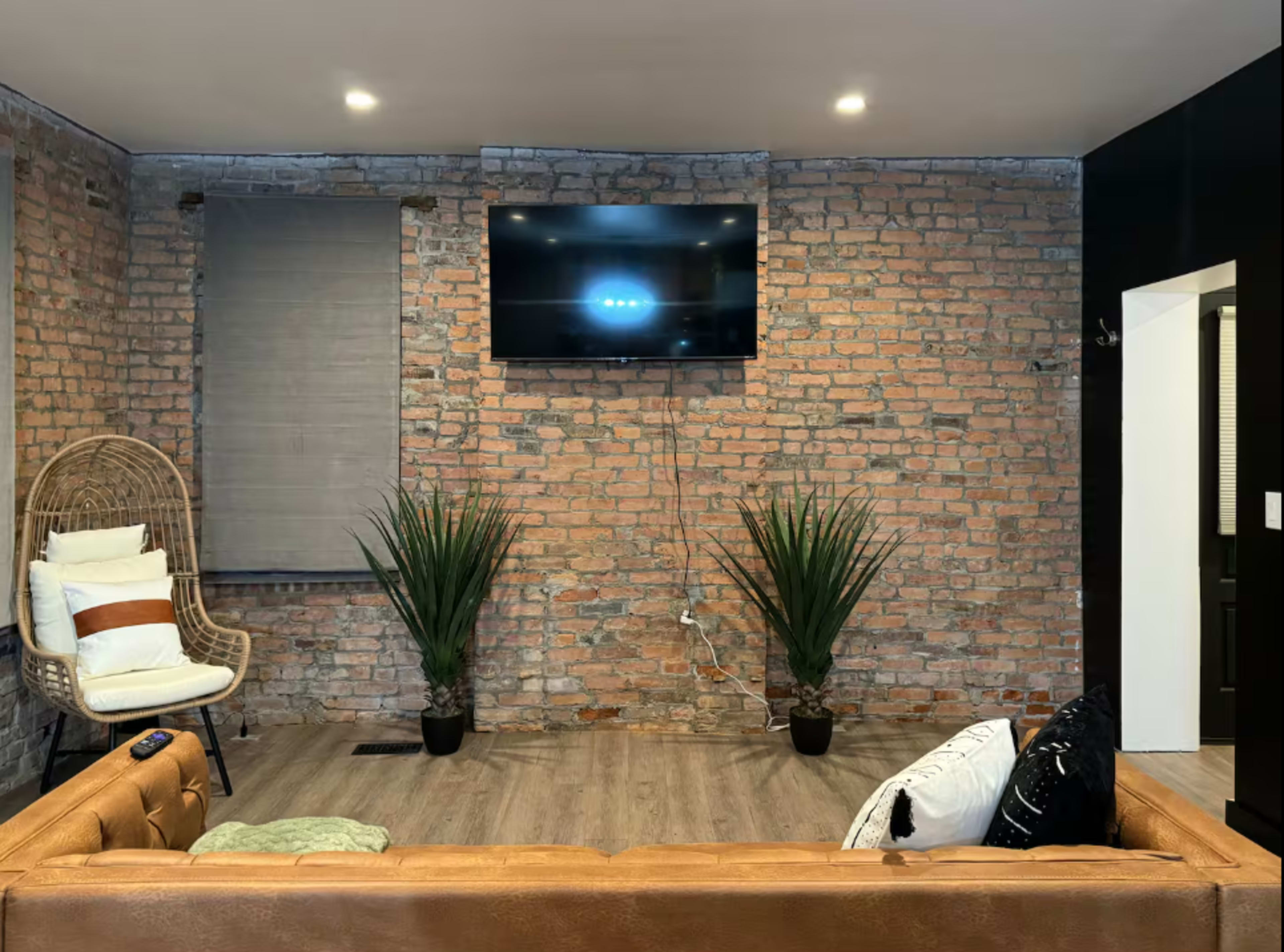 The image shows a living room with a brick accent wall, a brown leather sofa, a wall-mounted television, and two potted plants on the floor.