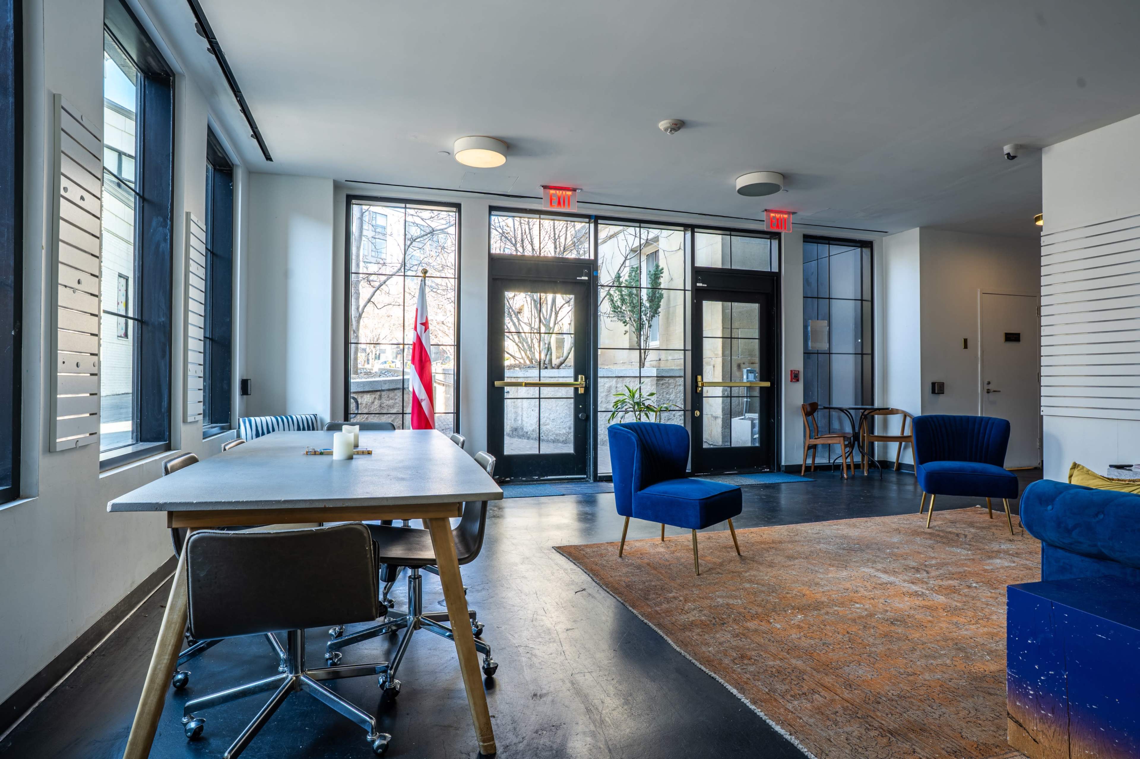 The interior of a modern space features a large table with an office chair, two blue armchairs, and a vintage rug, with large windows allowing natural light to enter.