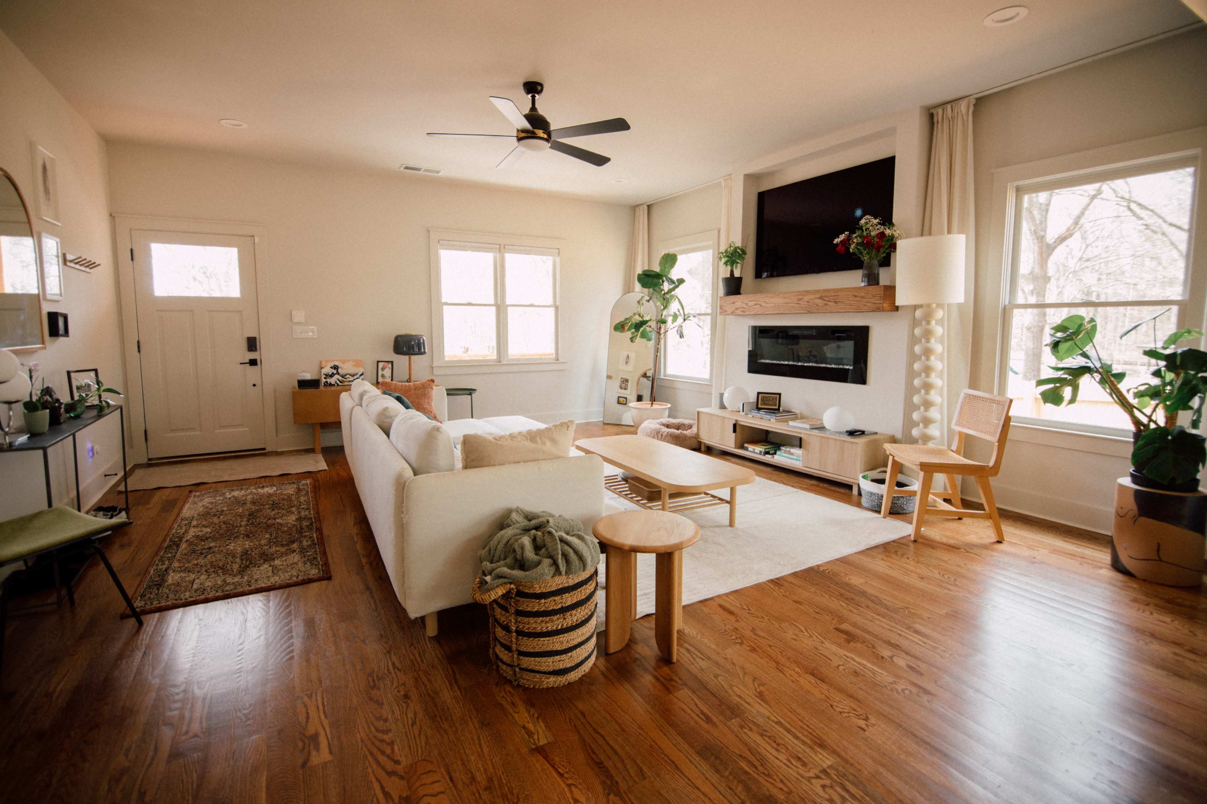 The spacious living room features a large white sofa, a wooden coffee table, and multiple plants, with natural light streaming through the windows.